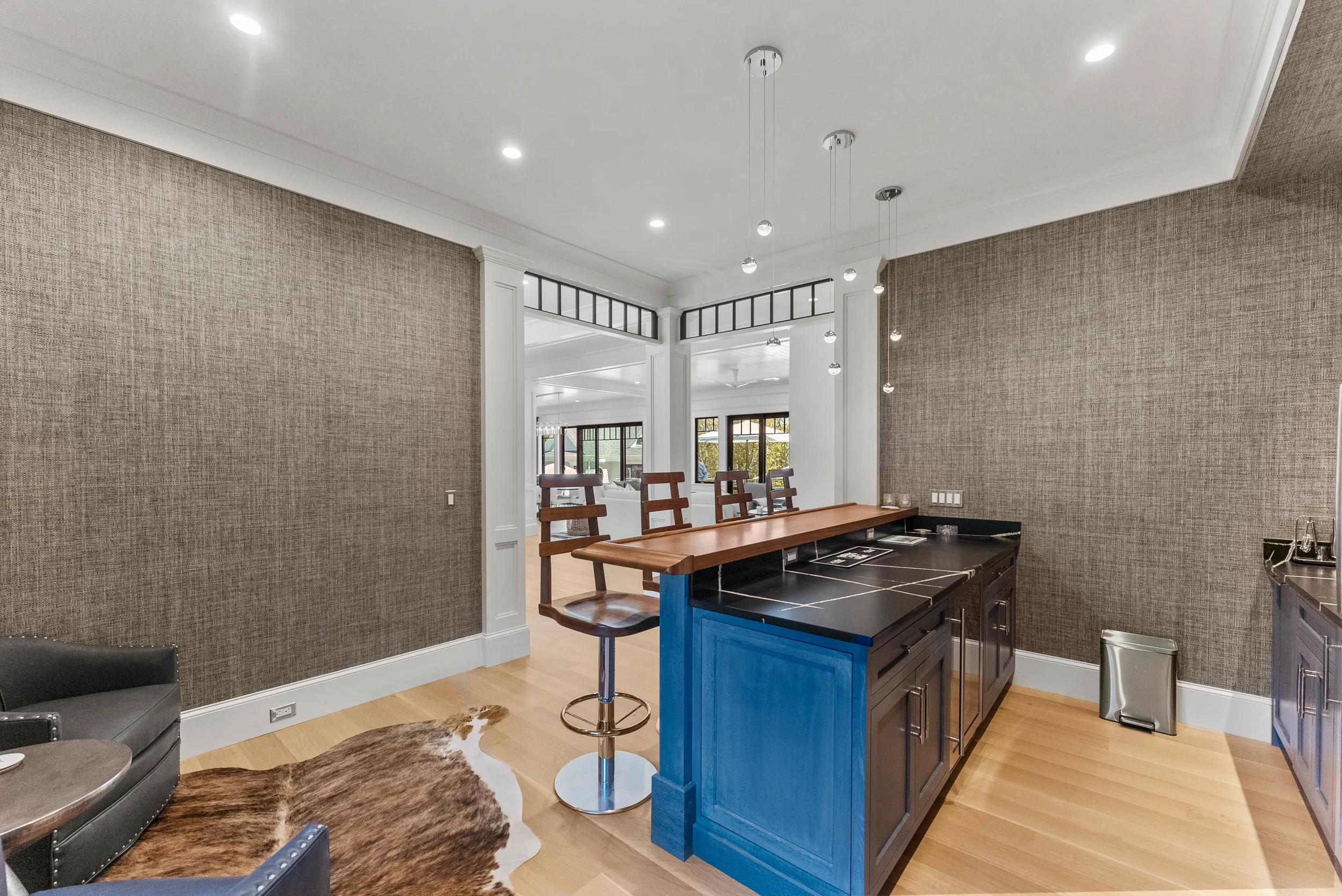 Interior of a modern kitchen and living area with a blue kitchen island, bar stools, a cowhide rug, and large windows with a view of a garden.