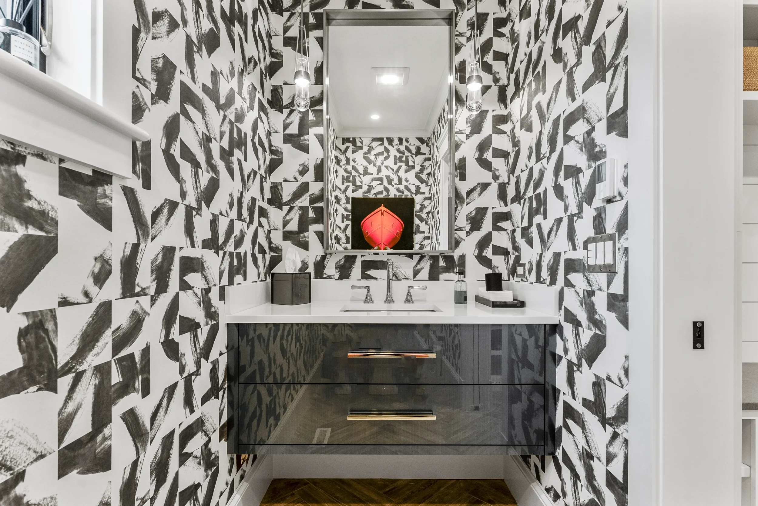 A modern bathroom with black, white, and gray patterned wallpaper. There is a white countertop with a sink, a large mirror, and black cabinetry underneath. Decorative items and toiletries are on the countertop, and a pair of hanging pendant lights il