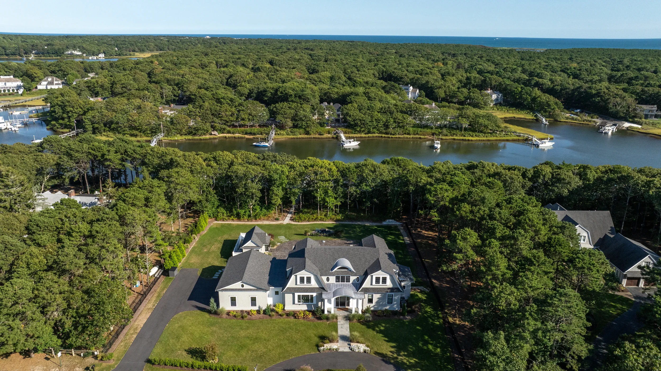An aerial view of a large white house with gray roof, surrounded by green trees, with a driveway leading to the street, and a backyard with lawn, near a waterway with boats, with dense forest and open skies in the background.