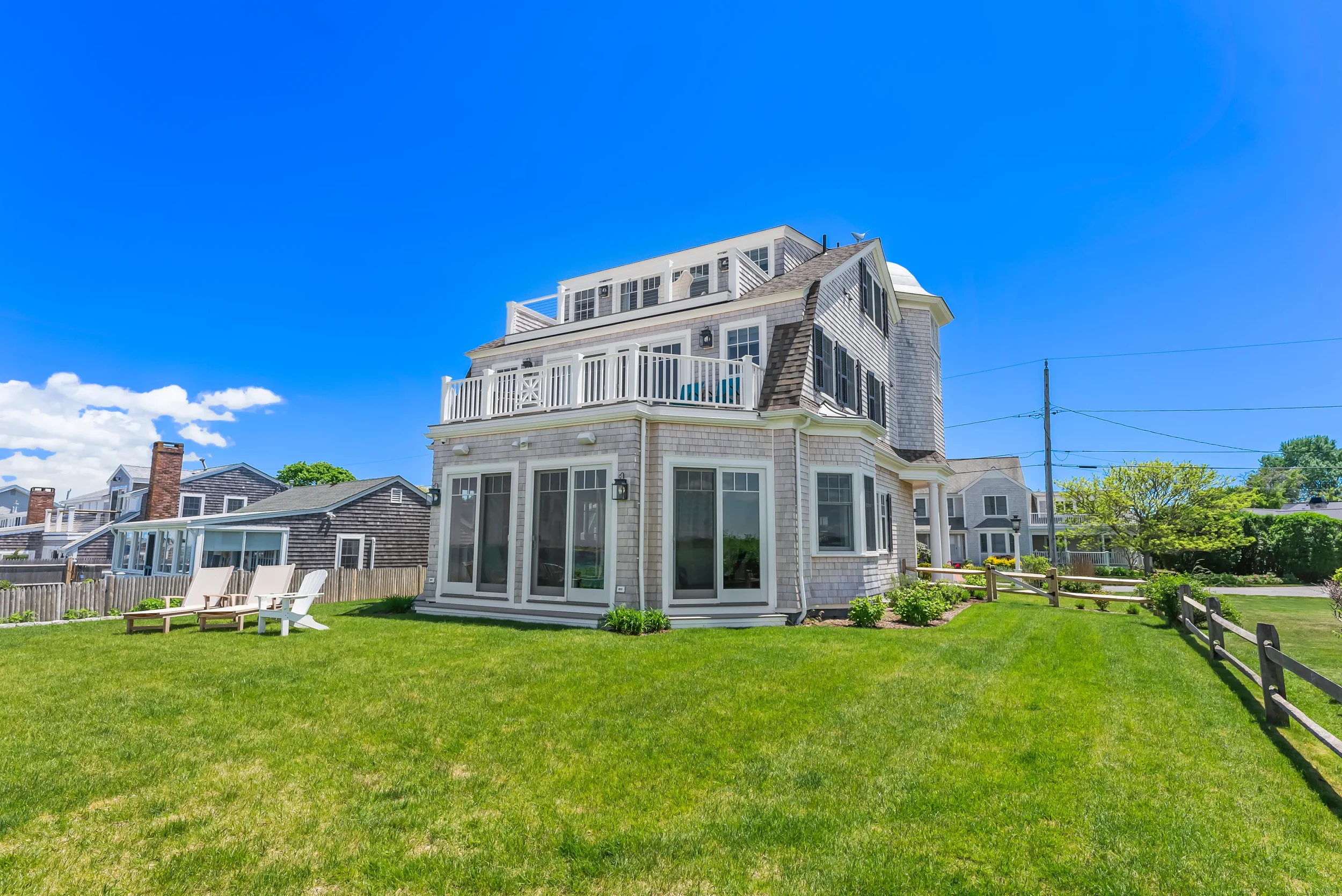 A large multi-story house with a gray exterior and multiple bay windows, sitting on a well-manicured lawn on a sunny day with a bright blue sky.