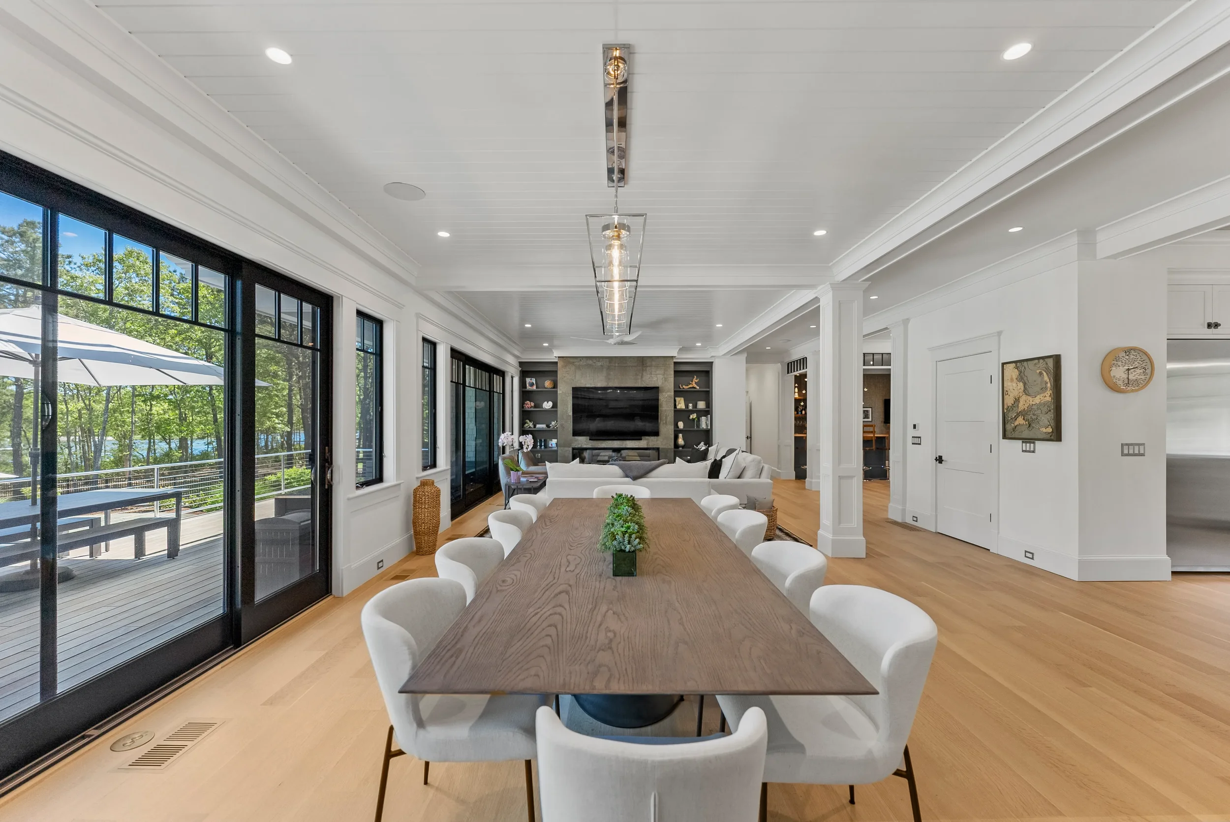 Spacious dining and living area with large windows, wooden floors, a long dining table with white chairs, and a living room with a gray sofa, TV, and built-in shelves.