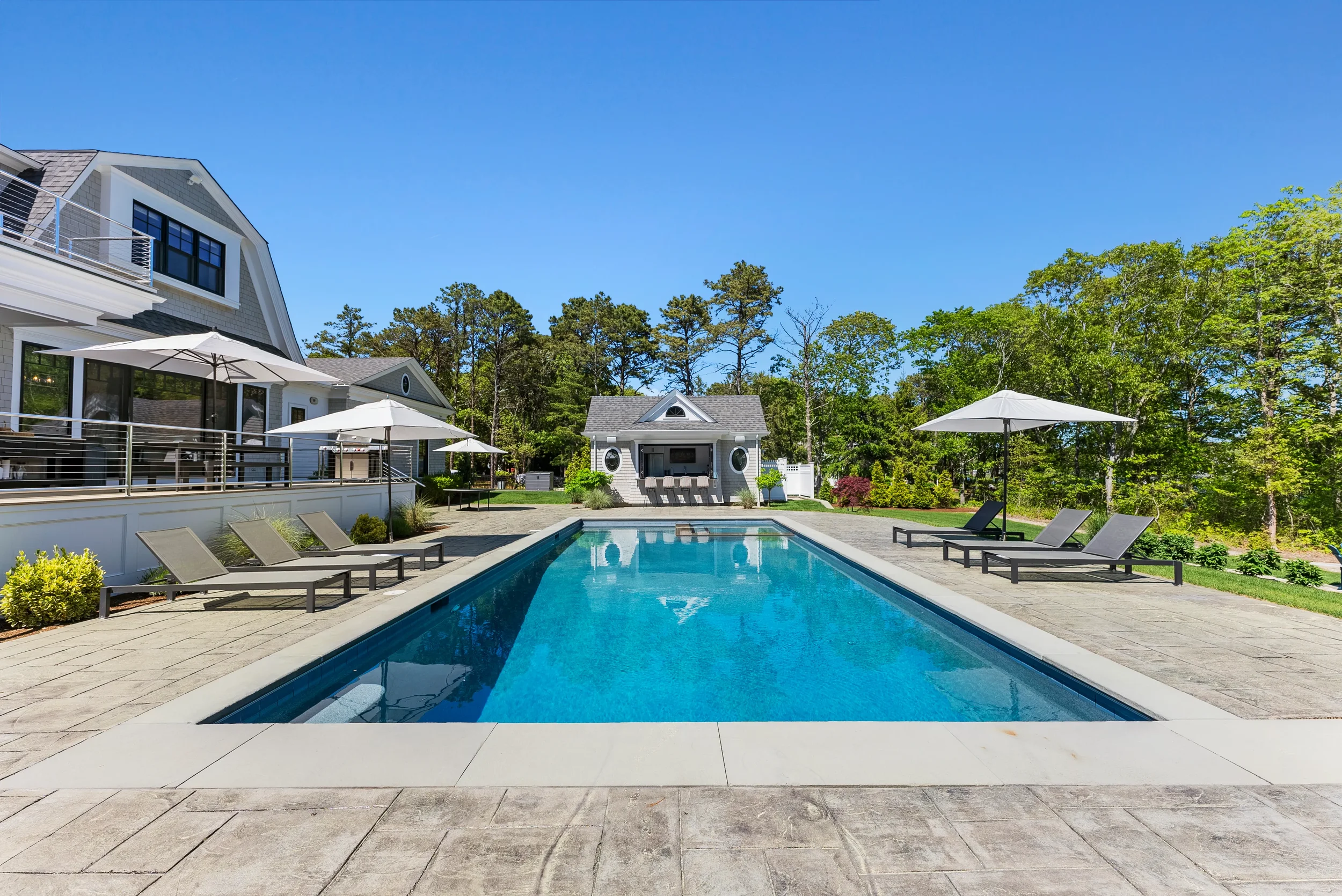 An outdoor swimming pool with lounge chairs and umbrellas, a small house in the background, surrounded by trees and a clear blue sky.