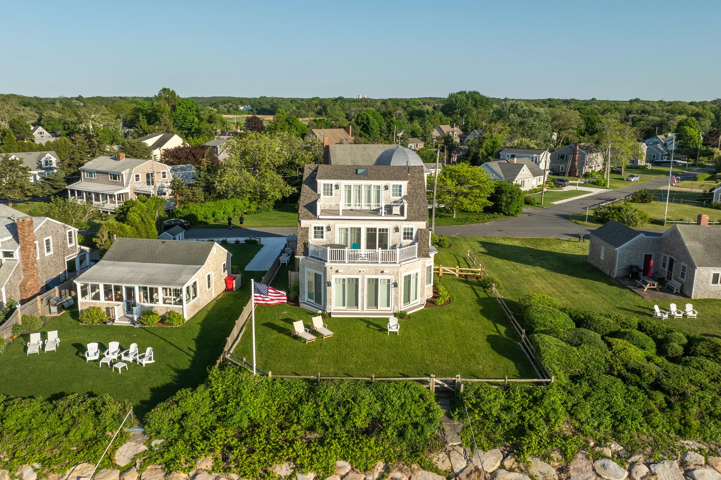 An aerial view of a large house with multiple decks, surrounded by a green lawn, American flag, and neighboring houses in a suburban neighborhood.
