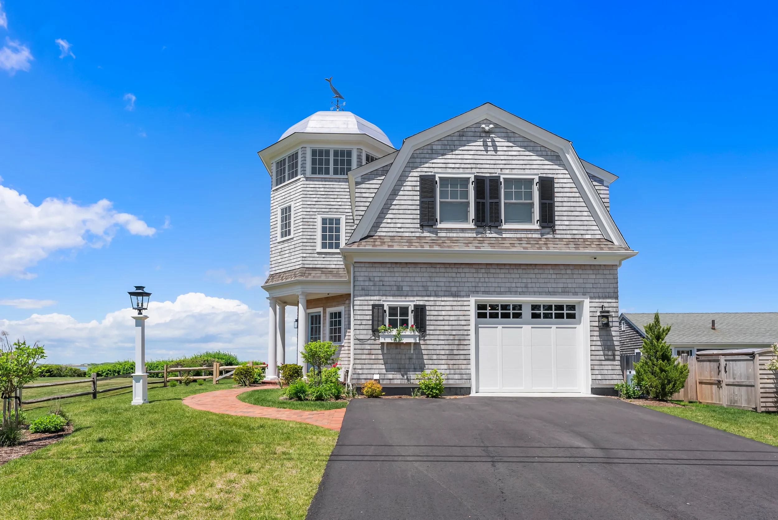 A two-story house with gray wooden shingles and black shutters, featuring a turret with a weathervane on top, surrounded by a landscaped yard with a brick walkway, streetlamp, green lawn, and a wooden fence under a blue sky with white clouds.