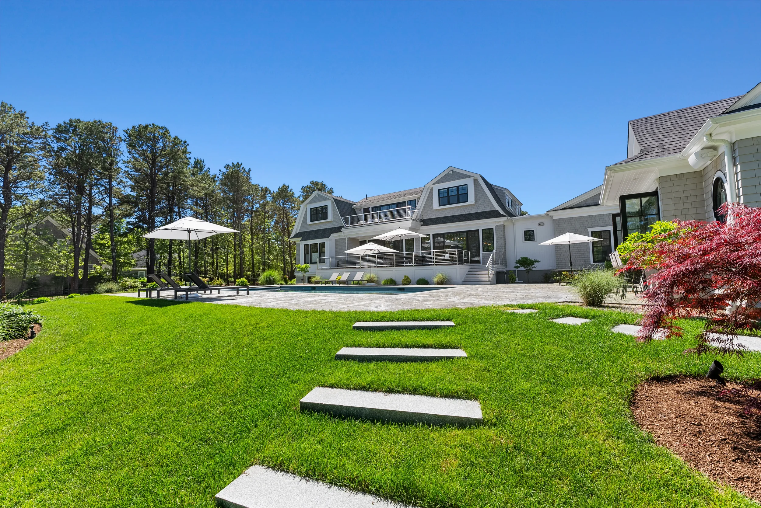 Beautiful backyard with green grass, stepping stones, outdoor lounge chairs, umbrellas, a swimming pool, and a large white house with black-framed windows and a deck, surrounded by trees on a sunny day.