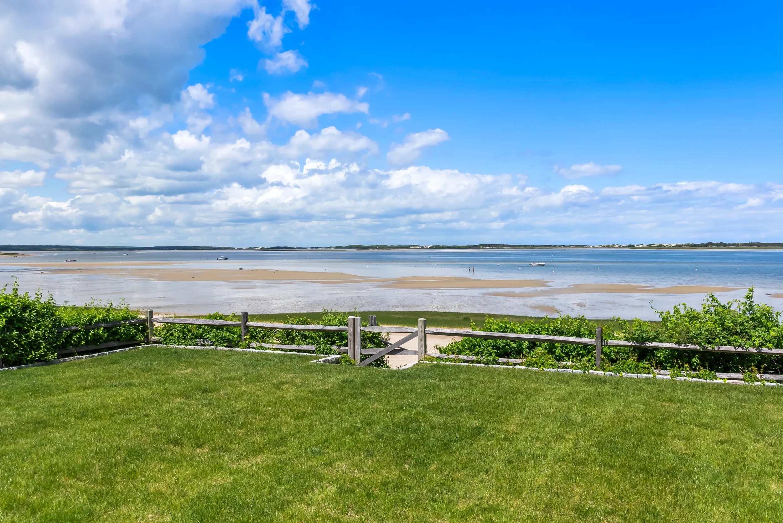 Scenic view of a grassy area with a wooden fence leading to a sandy and muddy shoreline, with calm water and boats in the distance under a partly cloudy blue sky.
