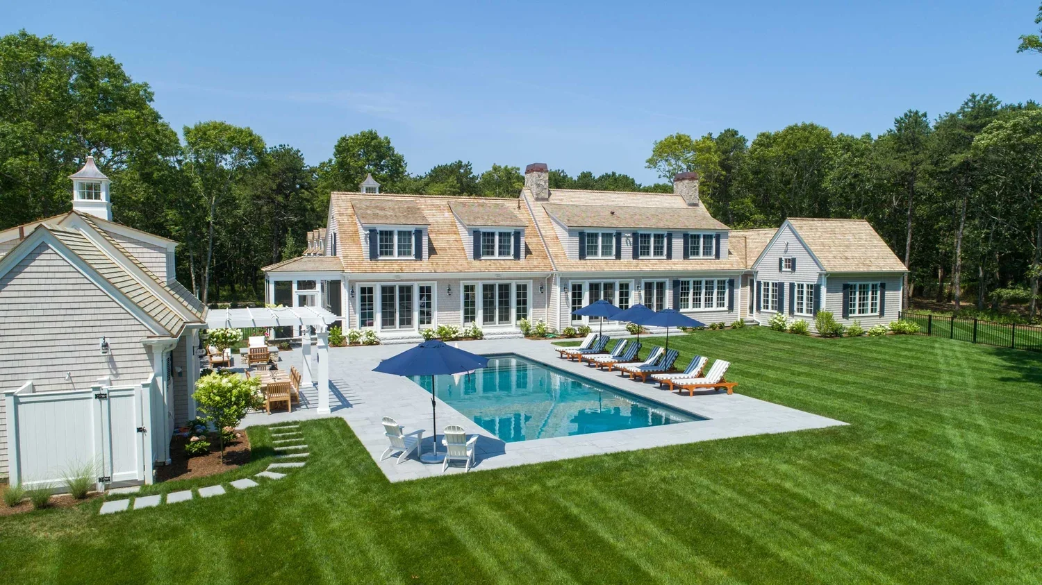 Large backyard with a swimming pool, surrounded by lawn and a white house with multiple windows and a patio with umbrellas and lounge chairs.