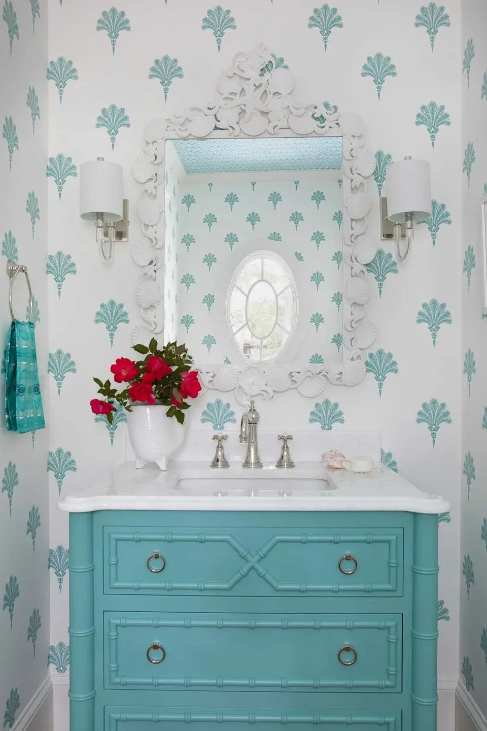 A bathroom vanity with a white marble top, turquoise cabinetry with decorative detailing, and a white sink with metal fixtures. Above it, a large mirror with an ornate white frame reflects a window with a decorative oval design. The walls are decorat
