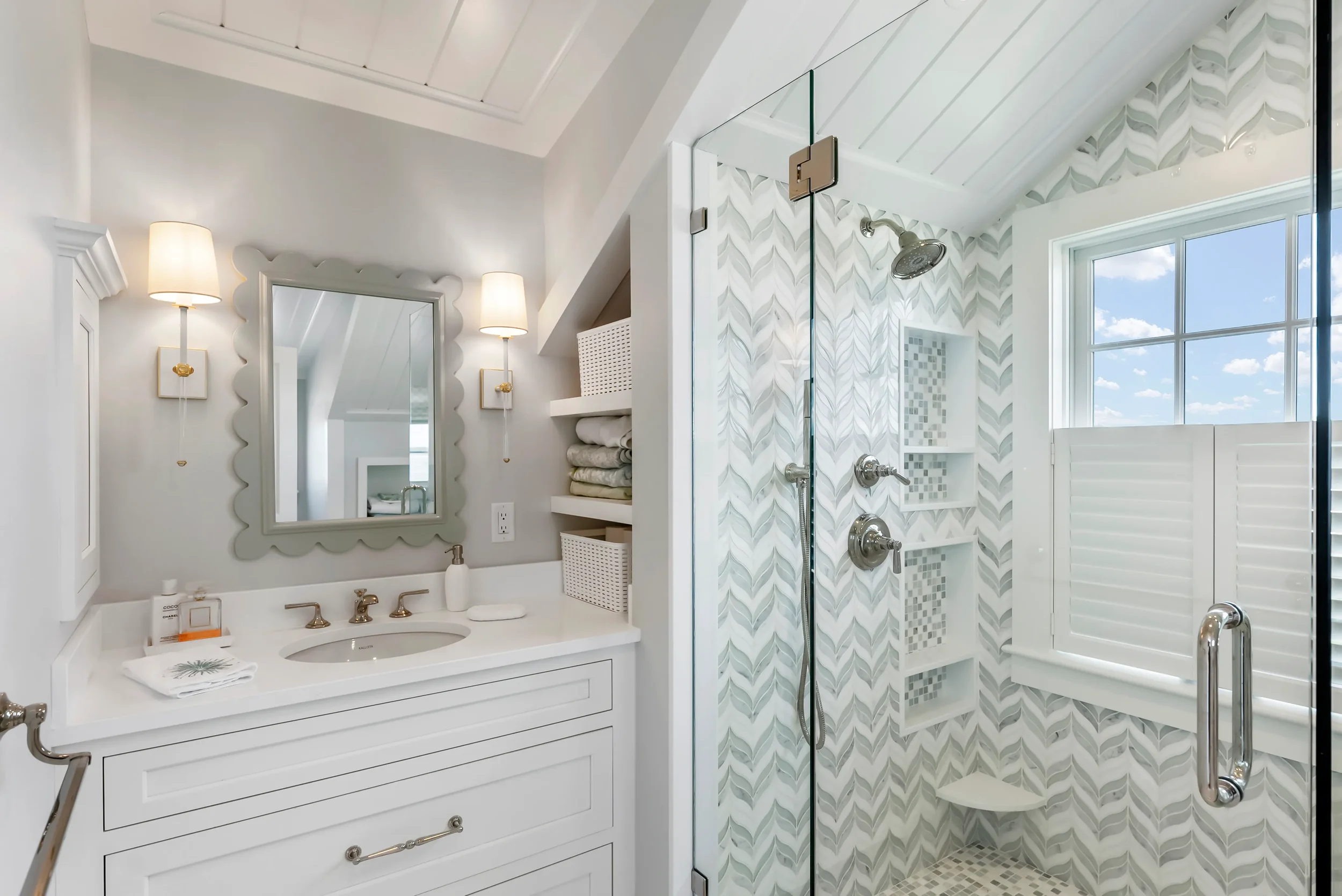 A bright, modern bathroom featuring a vanity with a mirror and wall-mounted sconces, a glass-enclosed shower with patterned tile, and a large window with white shutters.