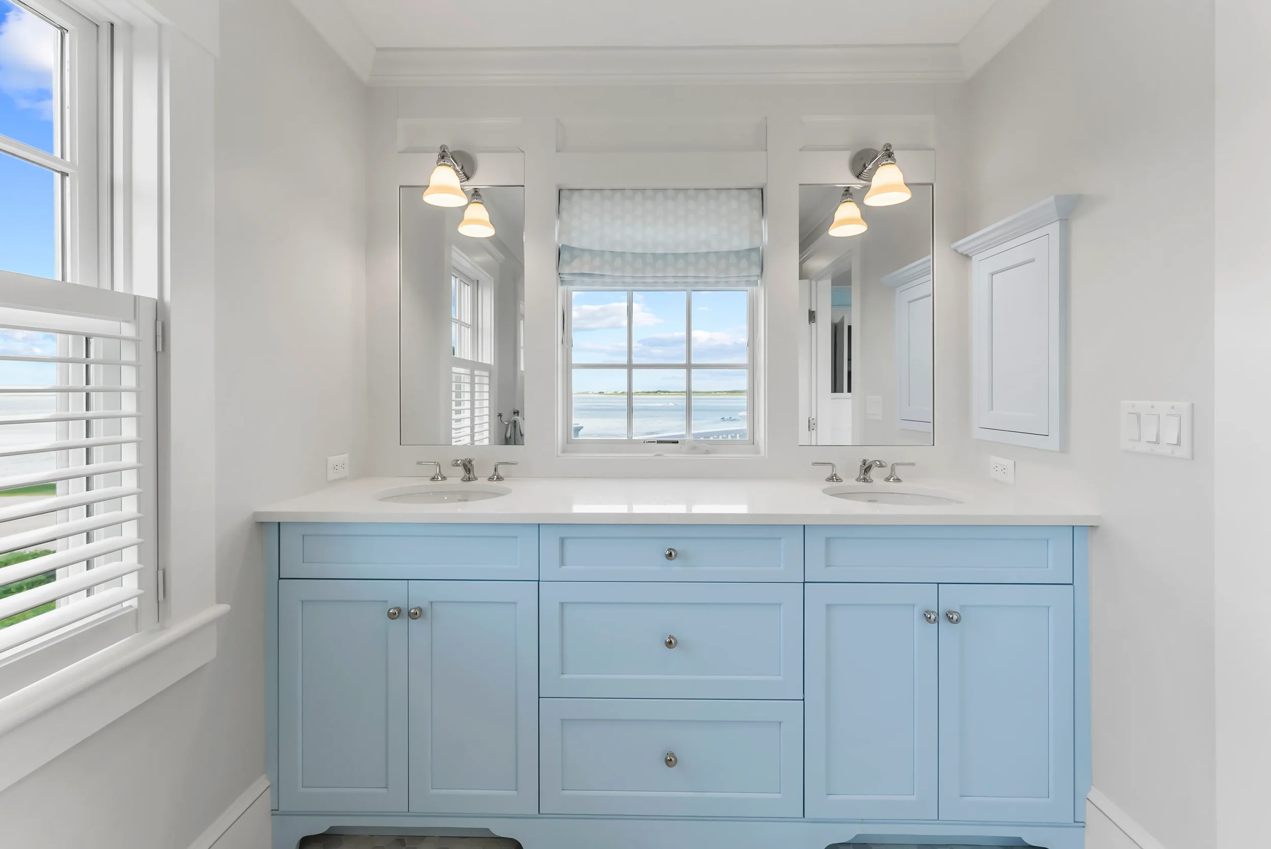 Bright bathroom with double vanity, light blue cabinets, white countertop, two mirrors, and a window with a view of a body of water