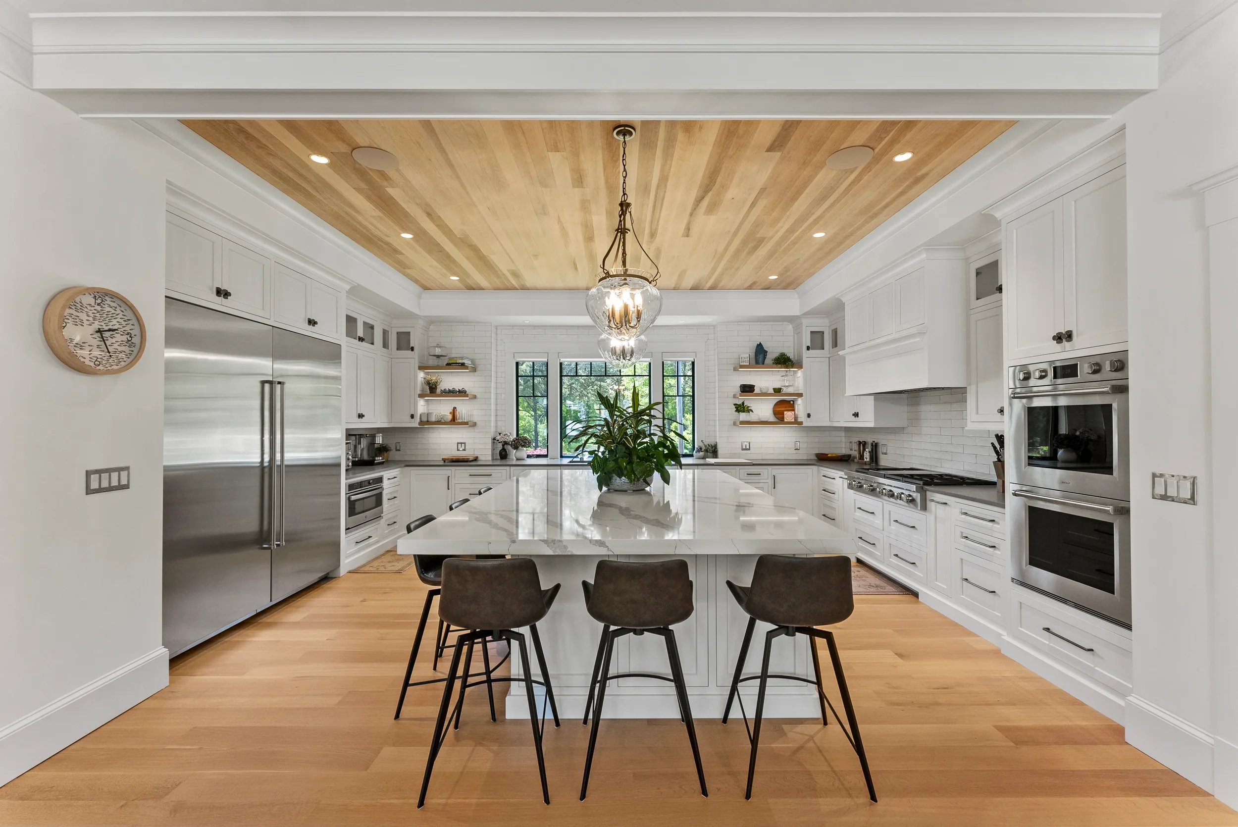 Modern white kitchen with a central island, stainless steel appliances, open shelving, hardwood floors, and a wooden ceiling with a hanging light fixture.