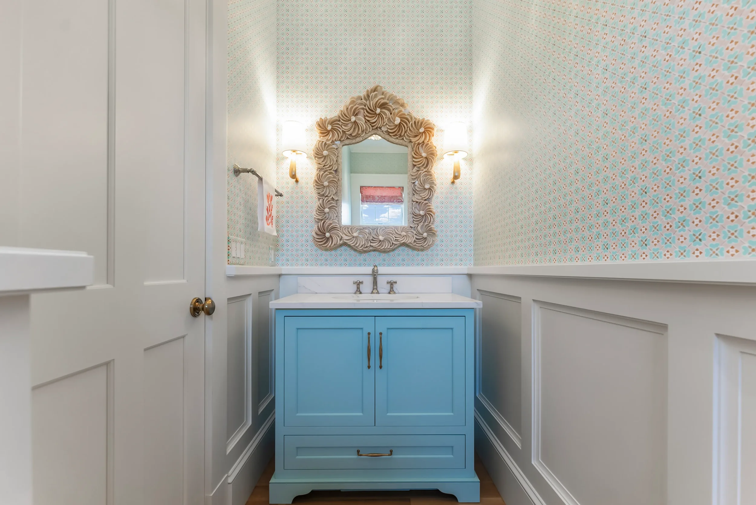 A small bathroom with a light blue vanity, a mirror with a decorative shell-shaped frame, wallpaper with pastel patterns, and wall sconces with white shades.
