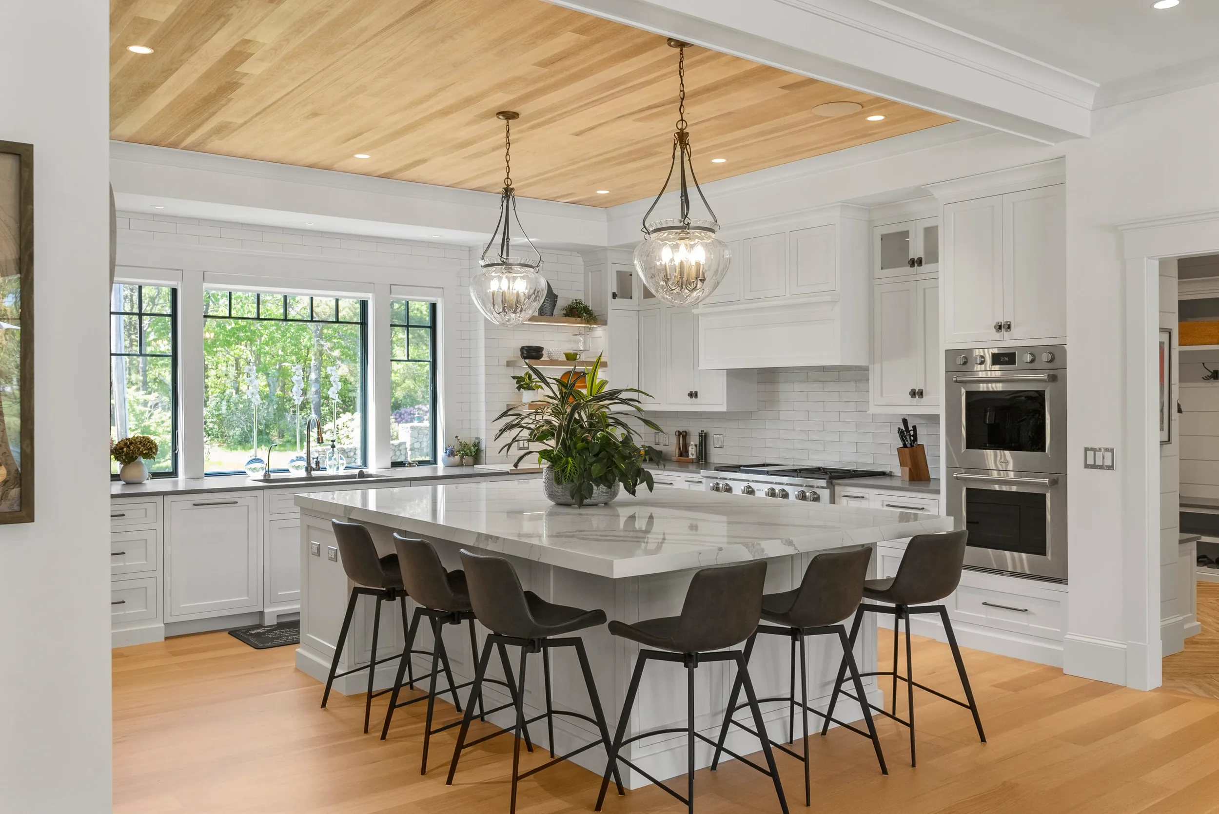 Modern white kitchen with large island, black bar stools, and pendant lighting. Hardwood floor and large window with outdoor view.