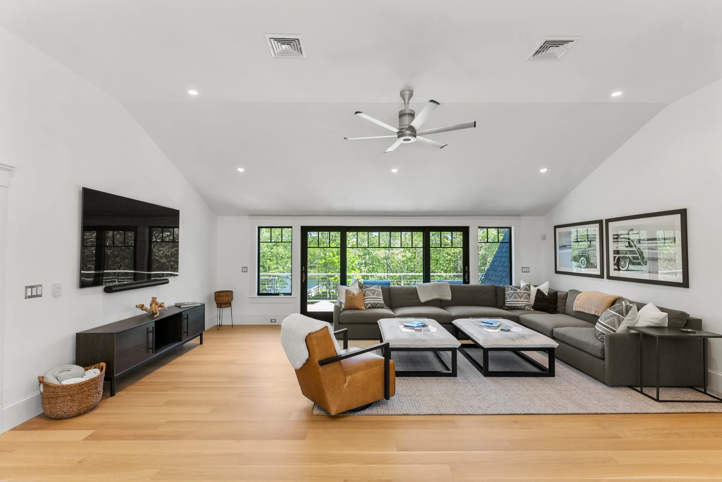 Modern living room with large beige sectional sofa, gray armchair with brown upholstery, wooden floor, black coffee tables, wall-mounted flat-screen TV, framed art, and large windows showing greenery outside.