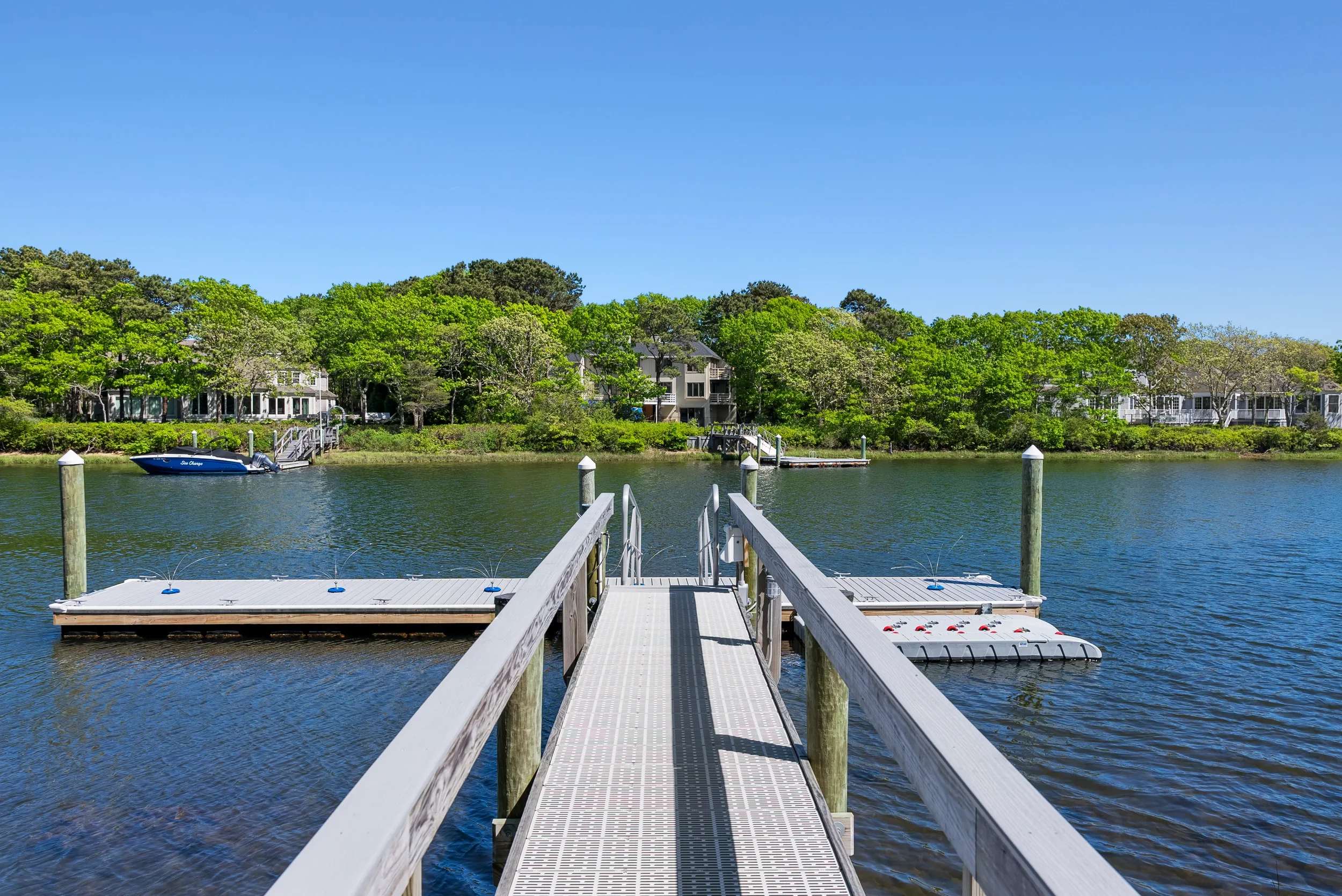 A dock extending into a calm body of water with a boat tied to it, surrounded by green trees and residential houses in the background under a clear blue sky.