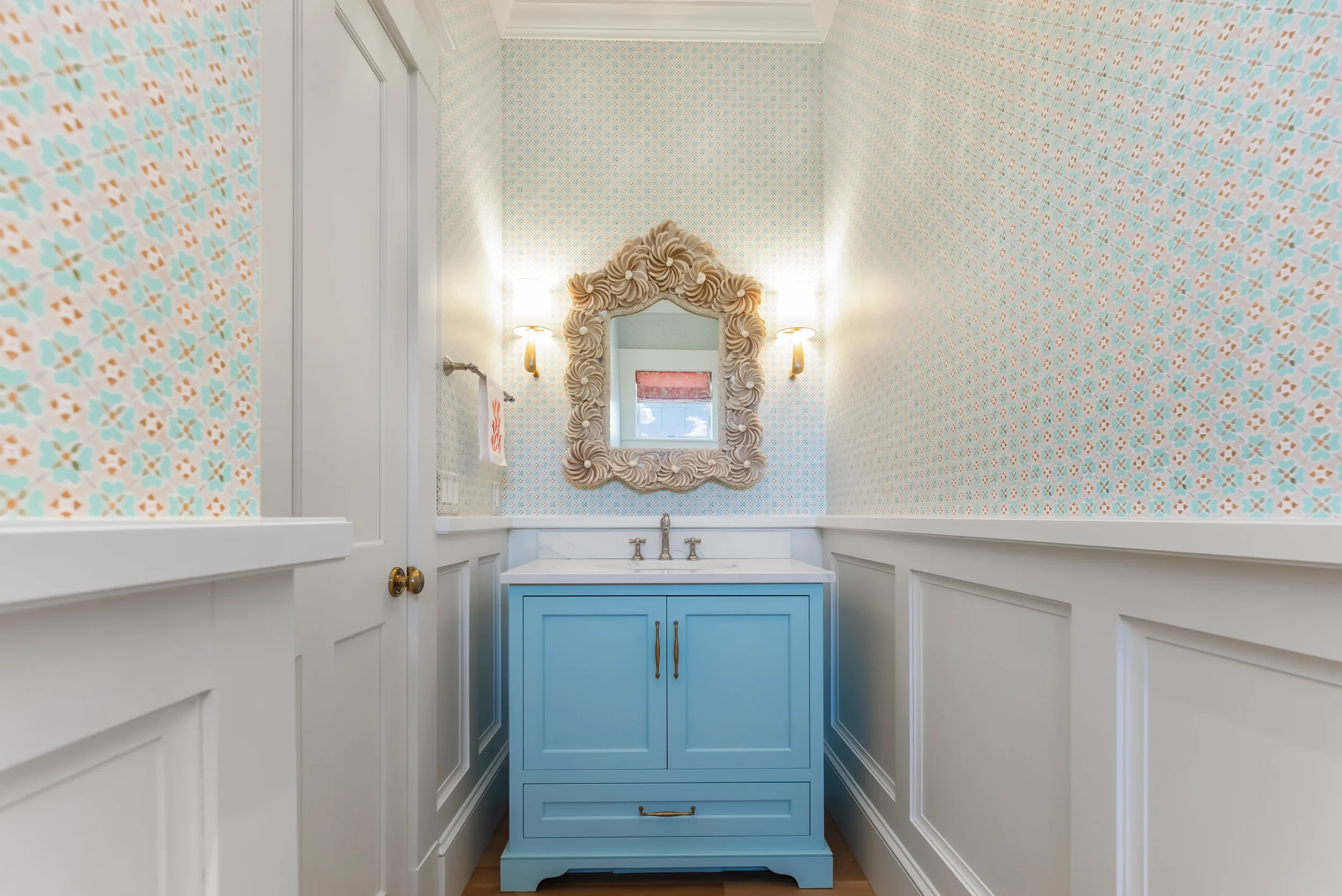 A small bathroom with patterned wallpaper, a blue vanity with a white countertop, a decorative mirror with a shell-like frame, and two wall-mounted lamps.