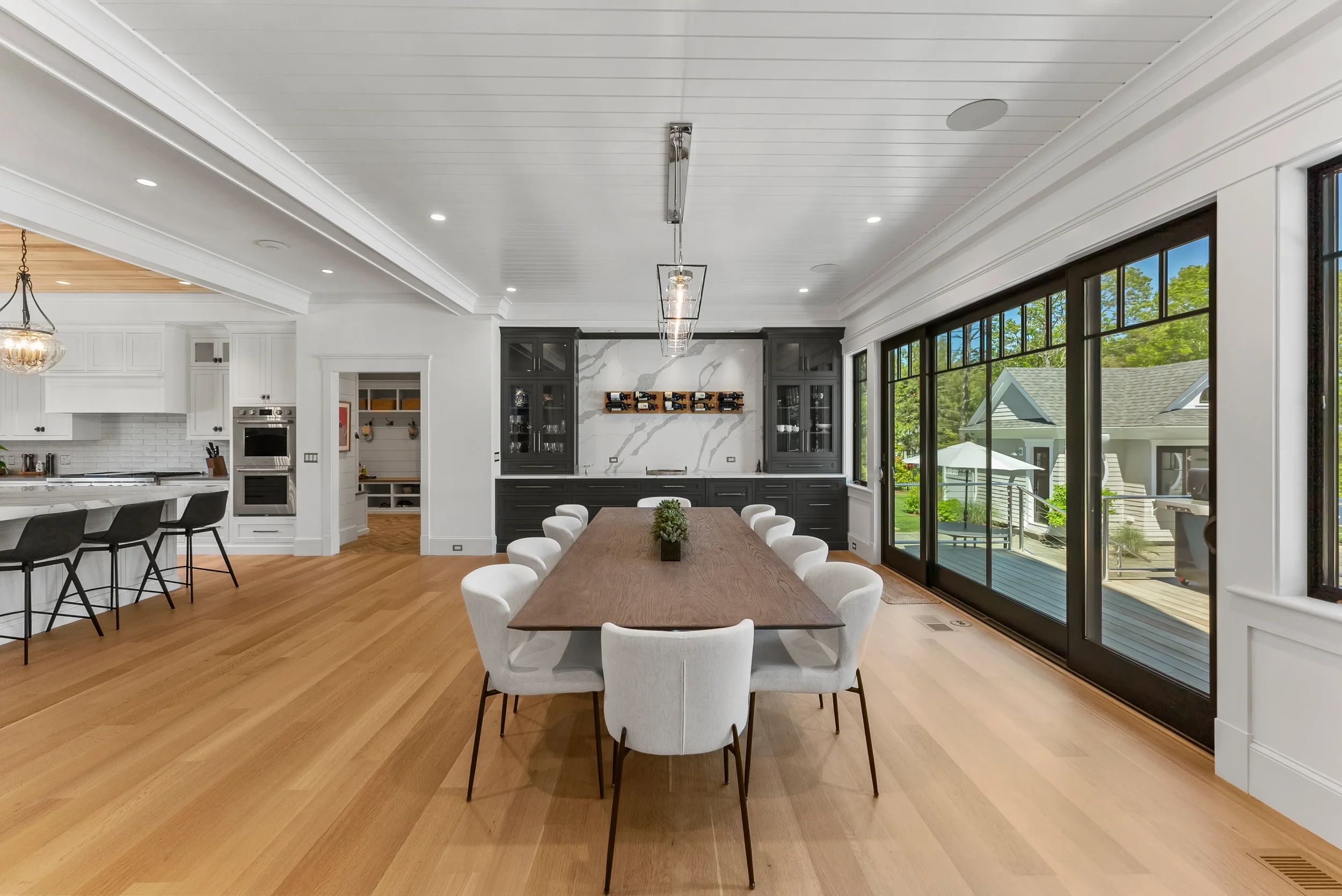 Modern dining room with large wooden table, white upholstered chairs, black cabinetry, marble backsplash, sliding glass doors to outdoor patio, and kitchen area with black bar stools.