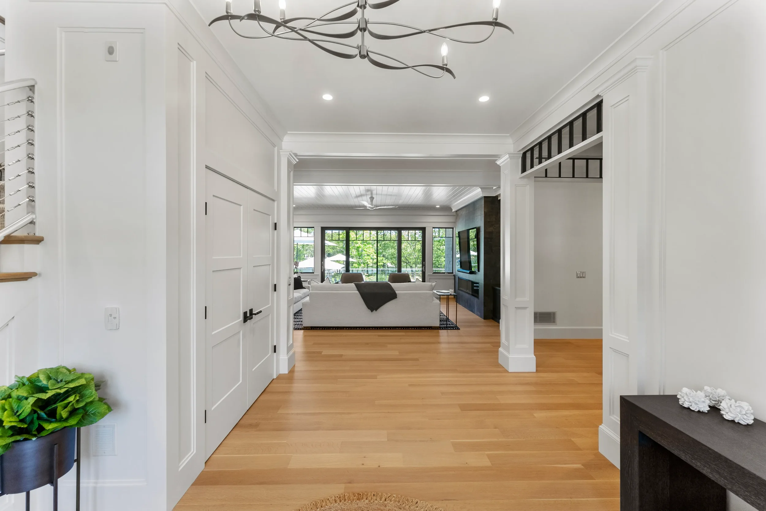 Living room with white walls, hardwood floor, modern chandelier, and large windows with greenery outside. Furniture includes a white sofa, TV mounted on dark accent wall, and small side tables.