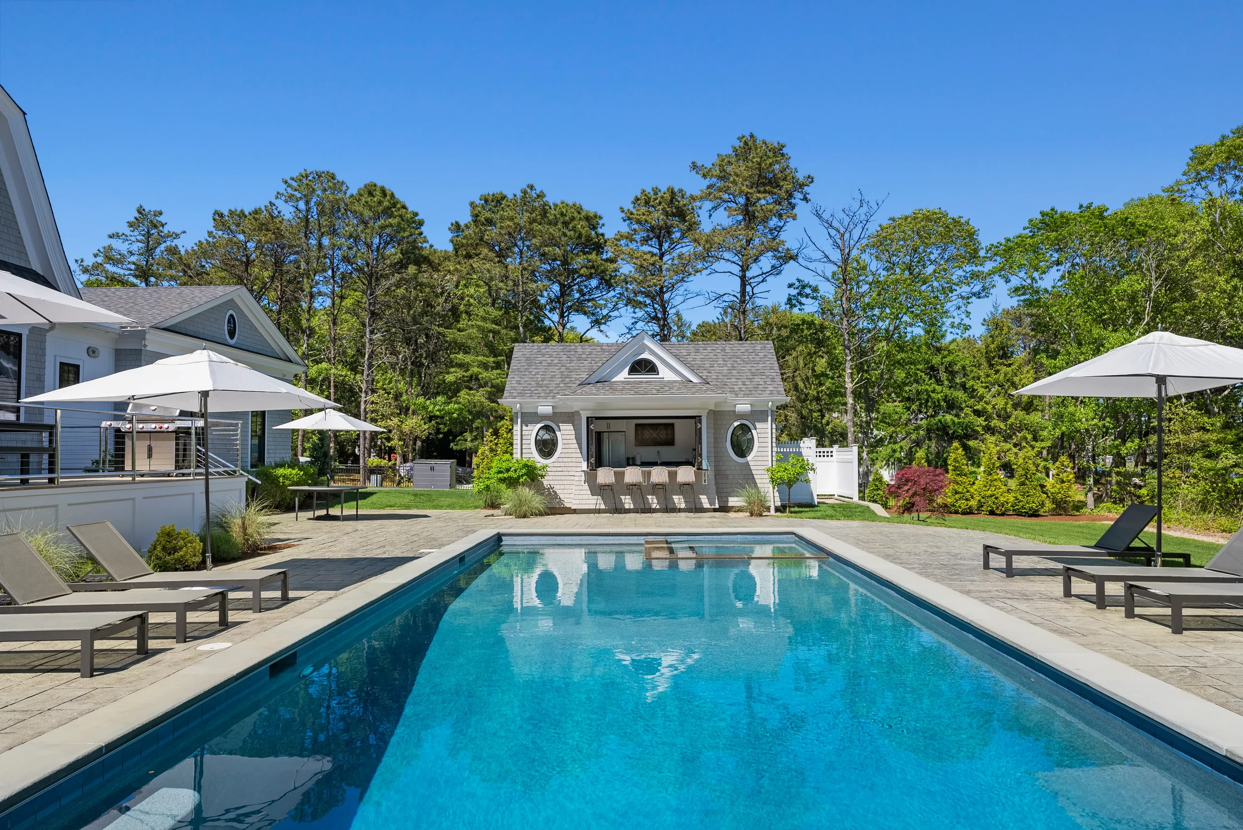 A backyard swimming pool with lounge chairs, umbrellas, and a poolside bar, surrounded by greenery and trees under a clear blue sky.