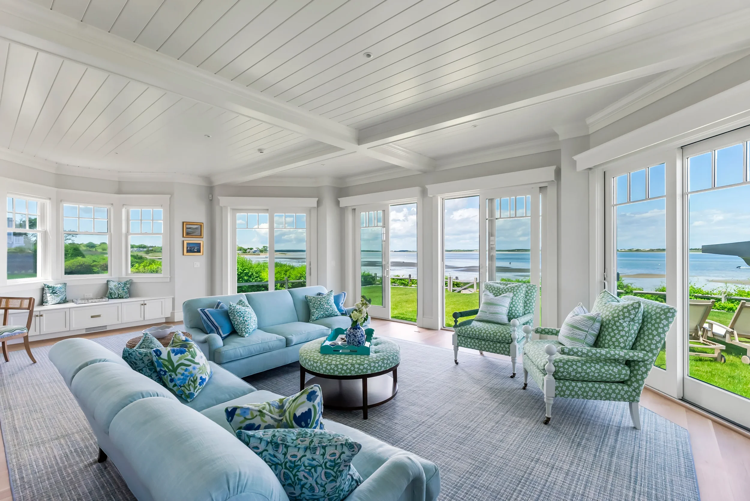 Bright living room with large windows overlooking a lake, featuring light blue sofas, green and white patterned armchairs, and a view of a grassy yard and water outside.