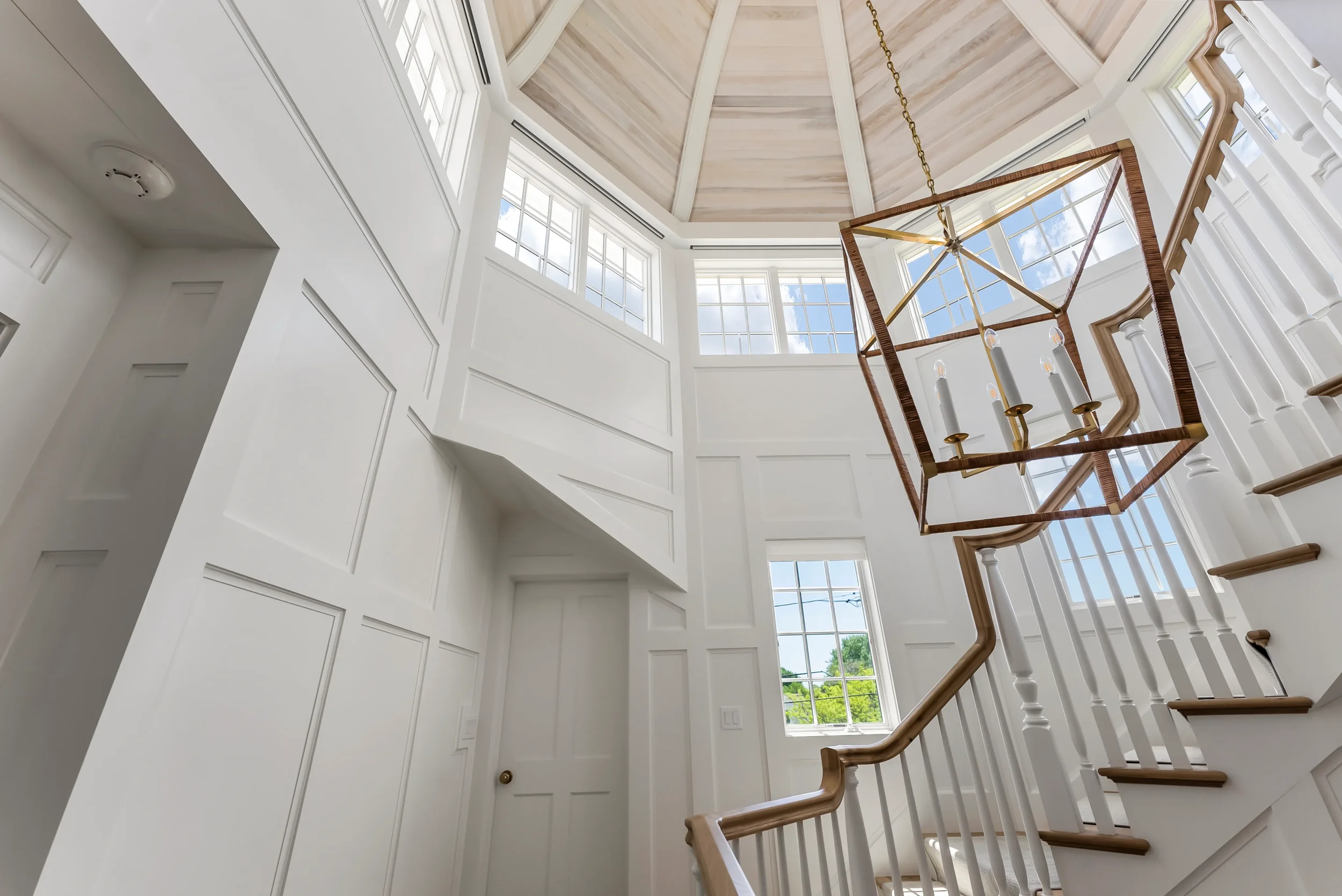 Interior view of a bright, two-story staircase with white walls, large windows, wooden handrails, and a modern geometric chandelier hanging from the ceiling.