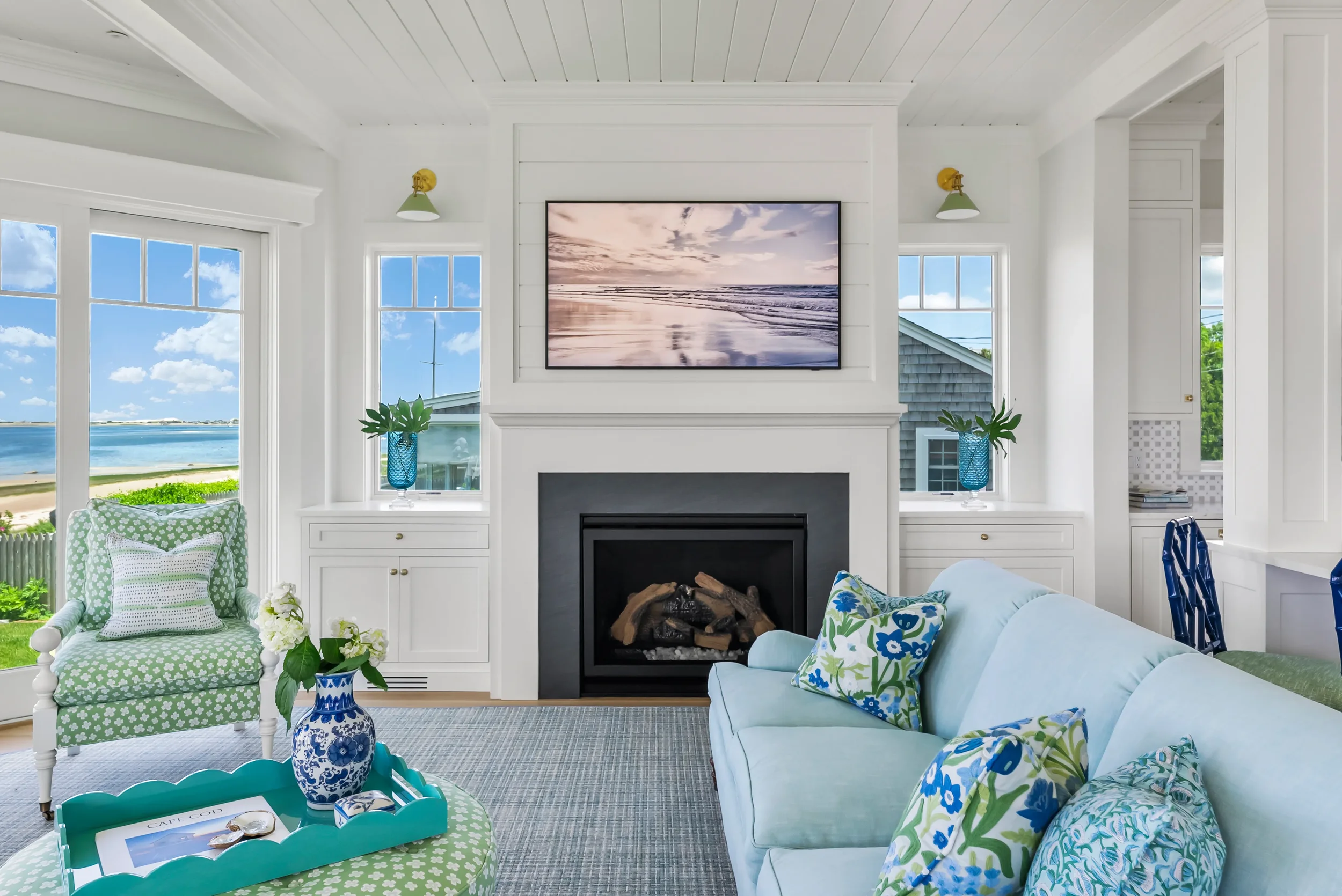 Bright living room with white walls and large windows overlooking a beach and ocean, featuring a fireplace with a television above, a light blue sofa with floral pillows, a green armchair with a patterned pillow, and a turquoise tray with flowers and