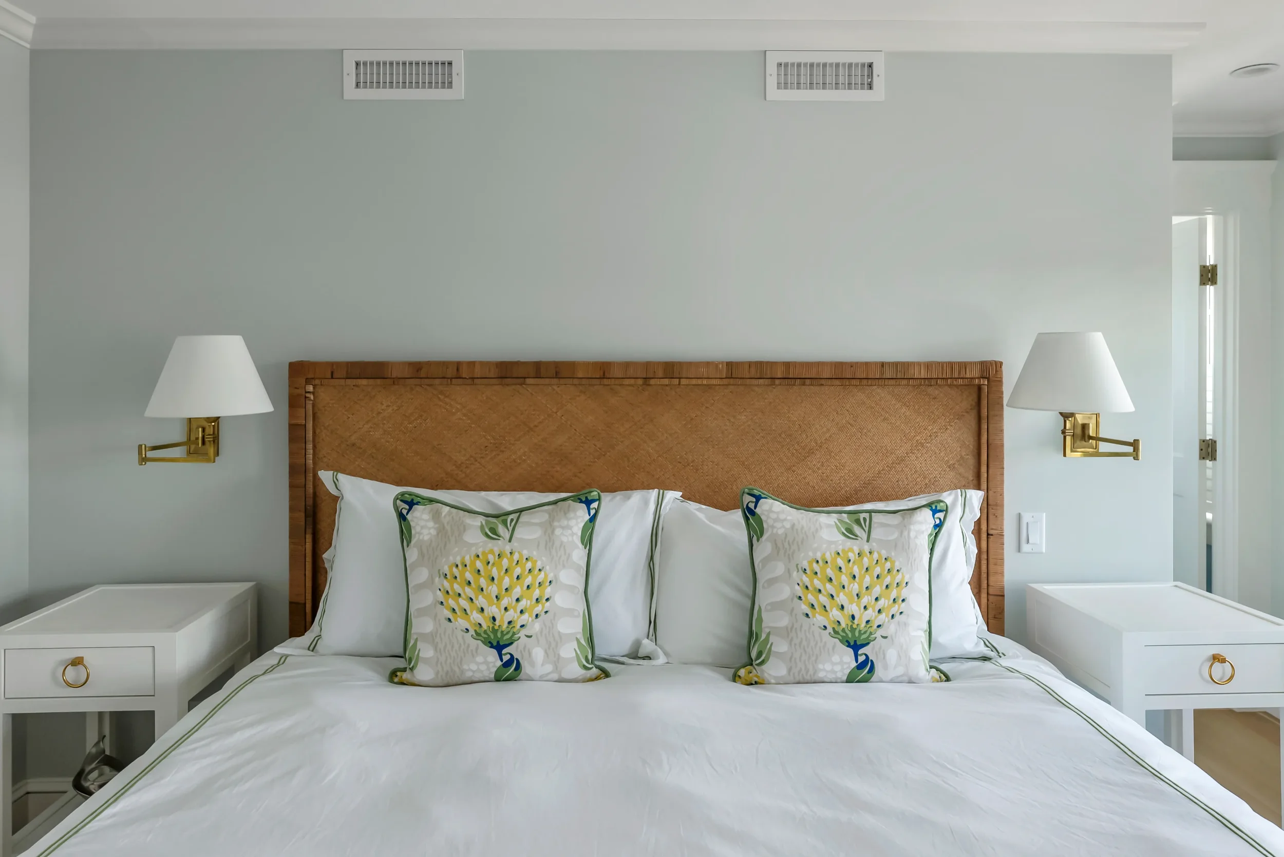 A neatly made bed with white bedding, three decorative pillows with yellow and green floral designs, a wooden headboard, and two white bedside tables with gold ring handles, each with a lamp attached to the wall above.