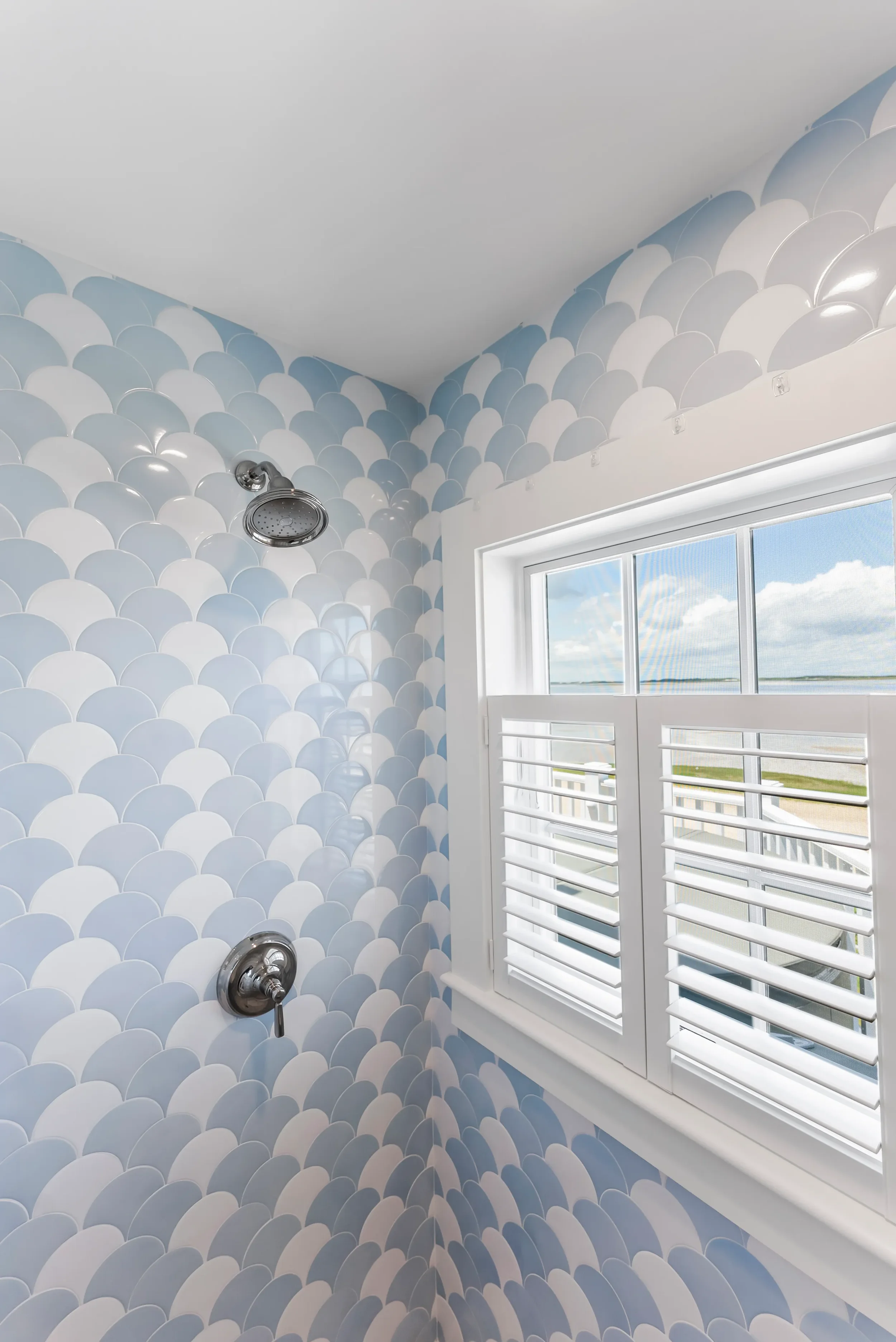 Shower with blue and white scalloped tile pattern, chrome showerhead, and white window with hurricane shutters showing a view of clouds and sky