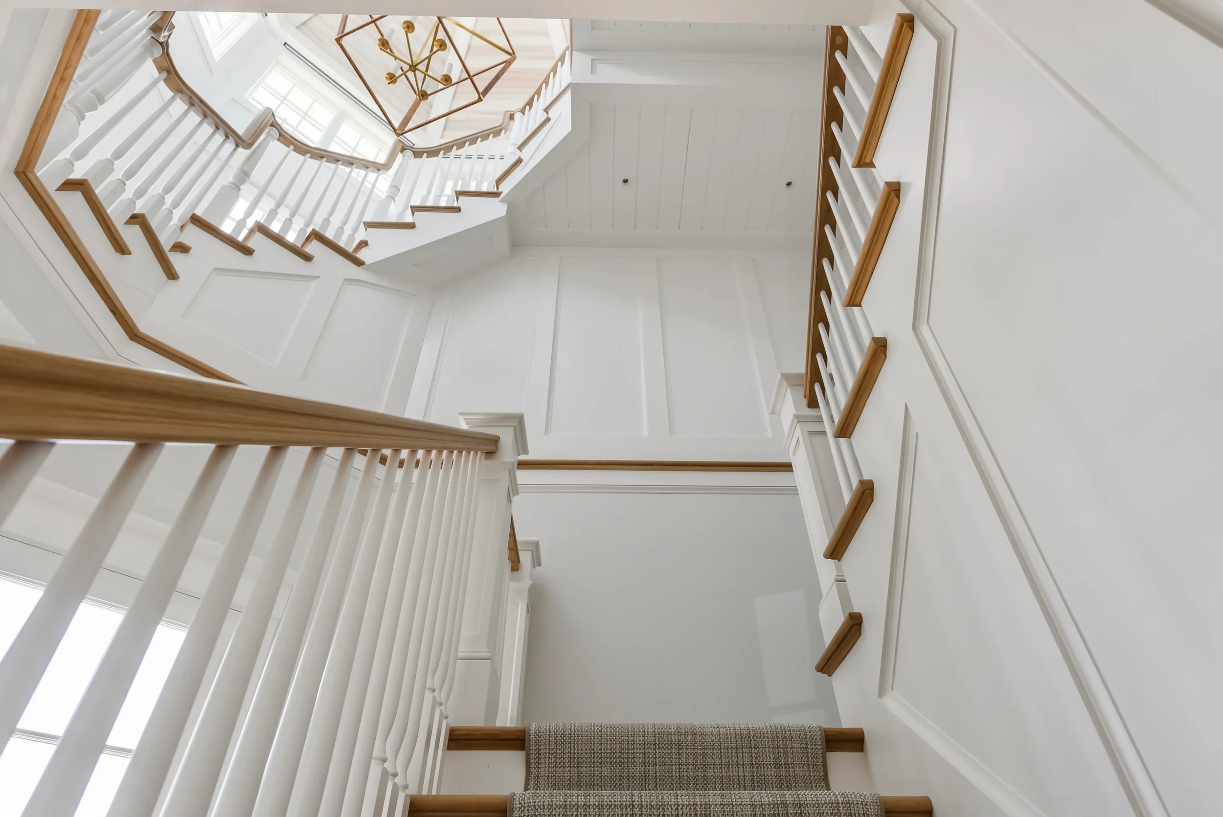 View looking up at a staircase with white walls, wooden handrails, white balusters, beige carpet on stairs, and a geometric chandelier hanging at the top.