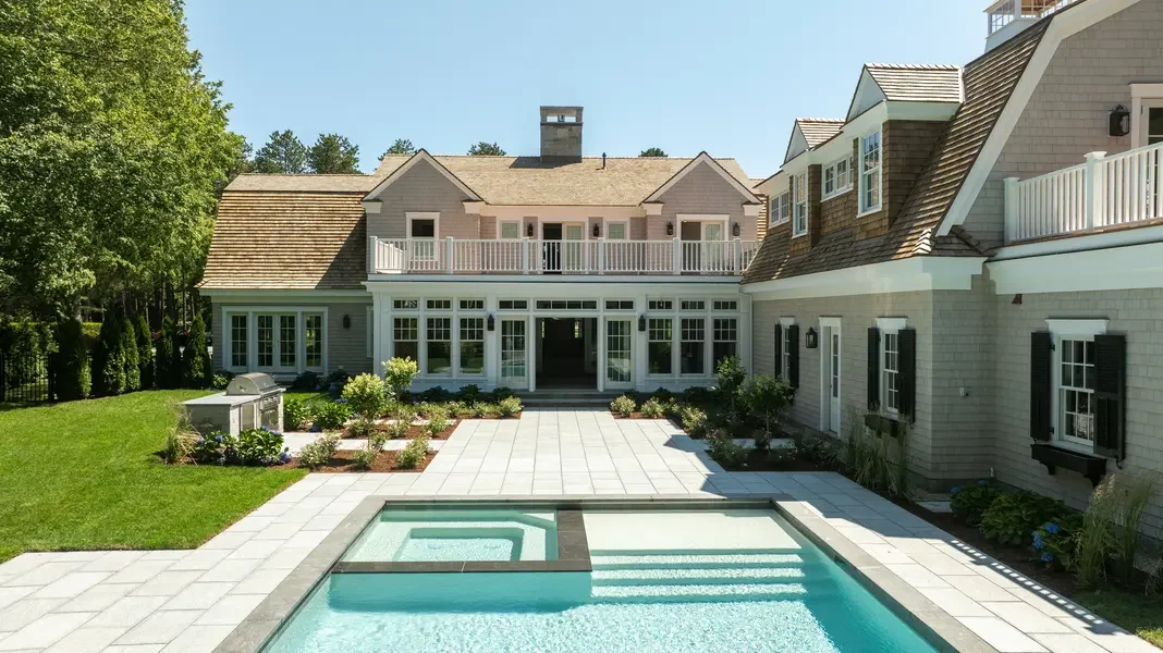 This image presents a magnificent luxury estate showcasing classic New England architecture arranged around a stunning pool courtyard. The expansive residence features weathered cedar shingle siding in soft gray tones complemented by crisp white trim