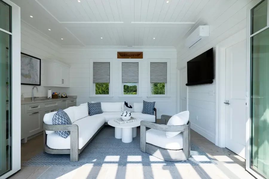 Cozy living room with a white sectional sofa, blue patterned pillows, a wood armchair with white cushions, a white oval coffee table, three windows with gray blinds, a wall-mounted TV, and a framed map on the wall.