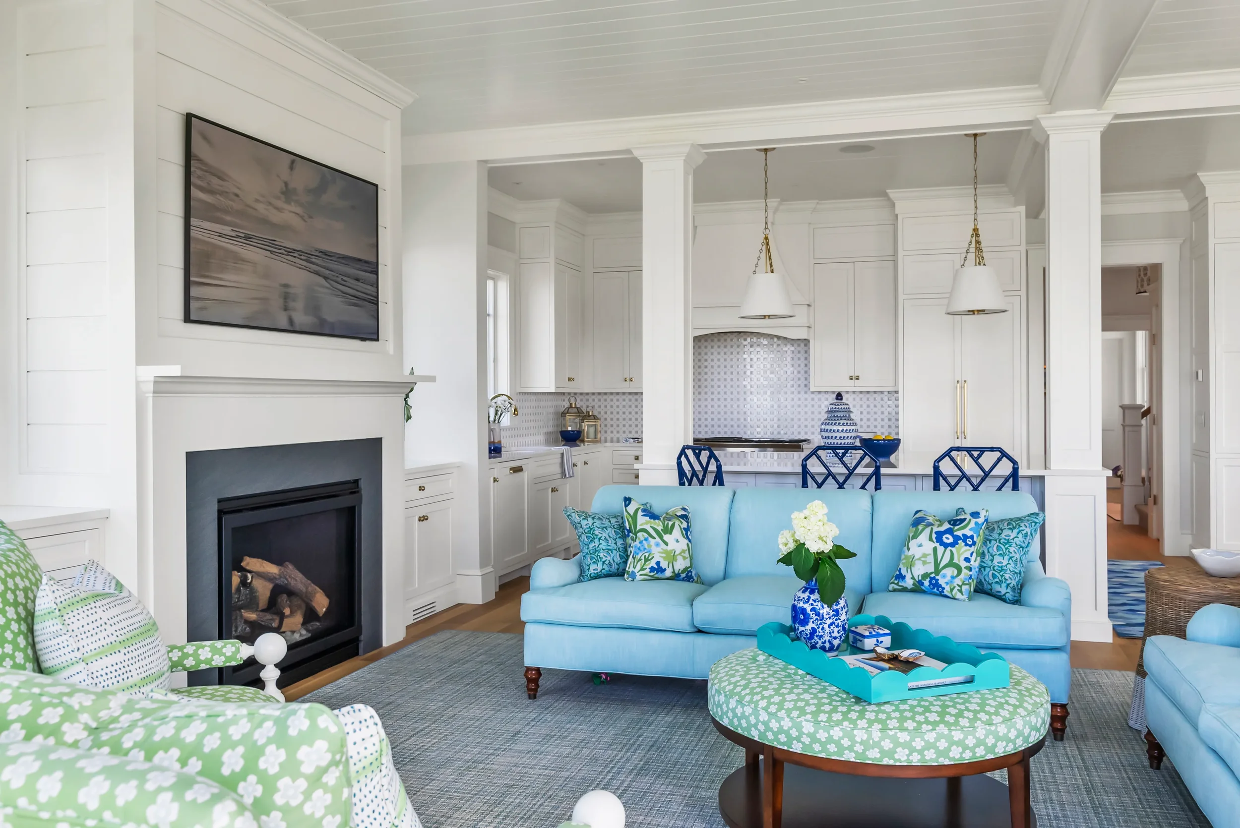 Bright, airy living room with white walls and cabinetry, a stone fireplace, a wall-mounted TV, a light blue sofa with patterned pillows, a round green ottoman with a tray holding decorative items, and a view into the white kitchen with pendant lights