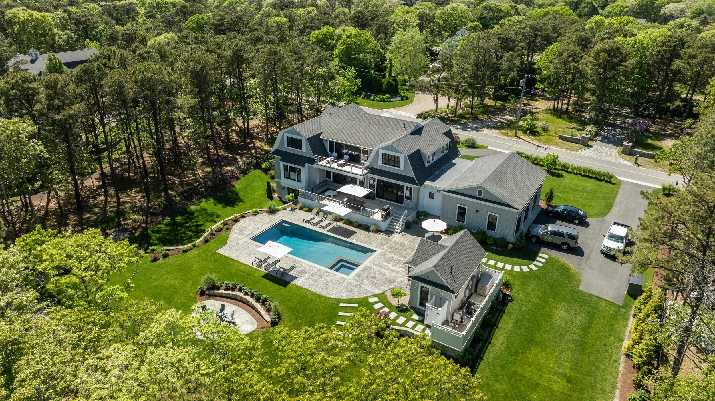 A large modern house with a backyard featuring a swimming pool, poolside lounge chairs, and a patio with umbrellas, surrounded by green lawns and trees.