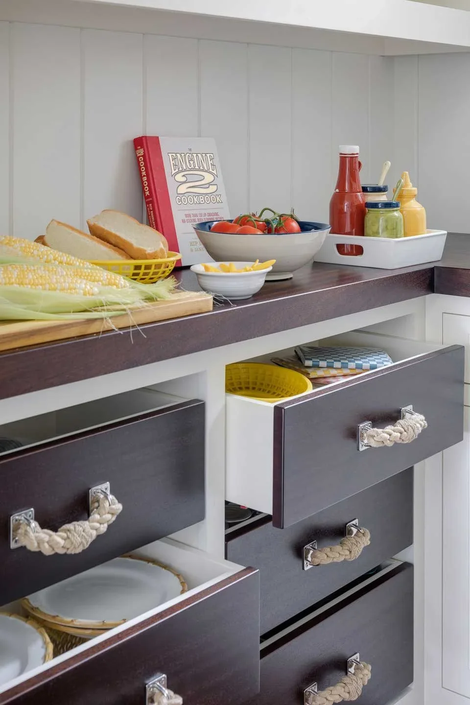 Kitchen countertop with bread, corn, tomatoes, condiments, and cooking books, partially opened cabinets with storage baskets and dishes.