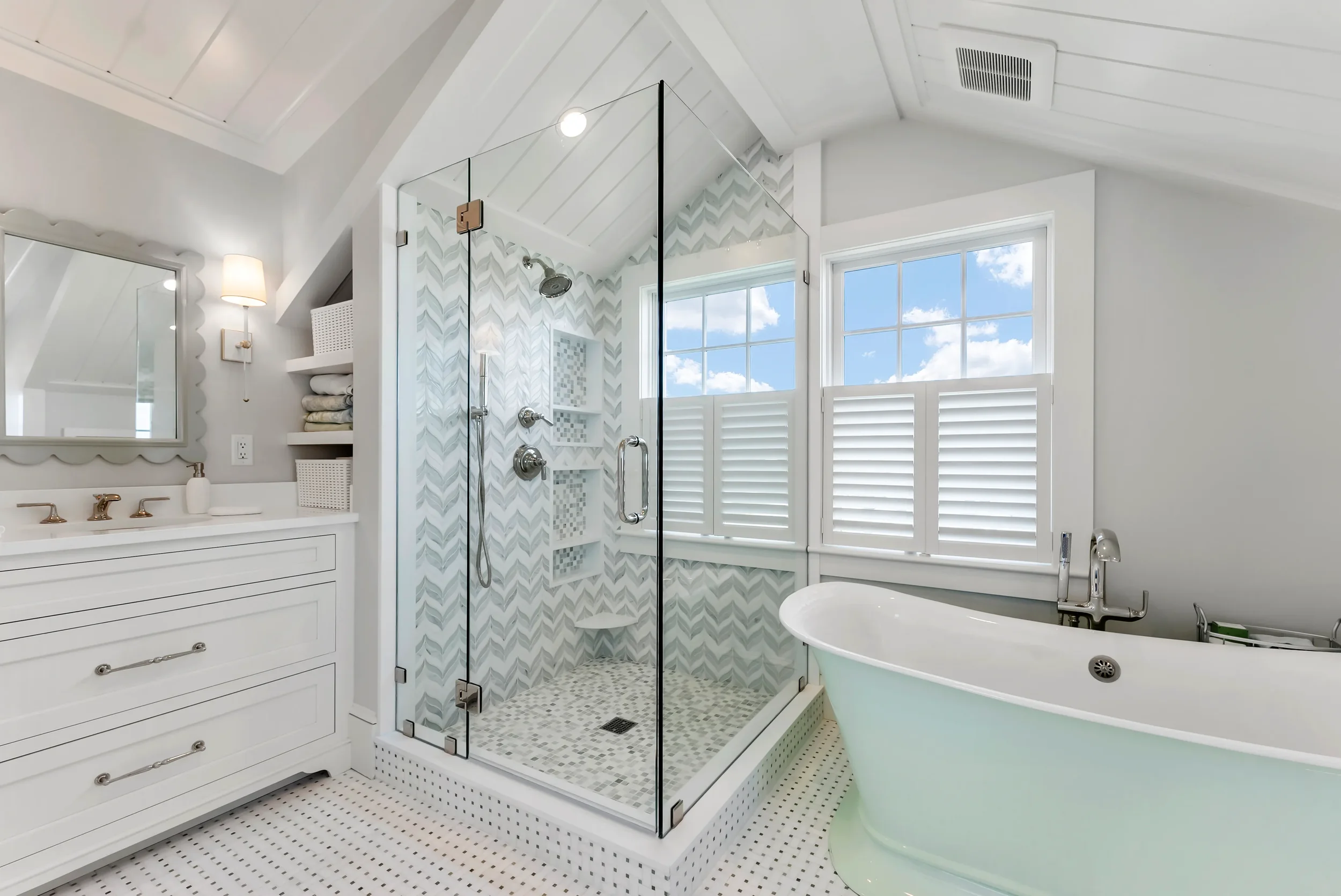 A bright, modern bathroom featuring a glass-enclosed shower with a patterned tile wall, a white freestanding bathtub, a vanity with a mirror and gold fixtures, and large windows with white shutters allowing natural light.