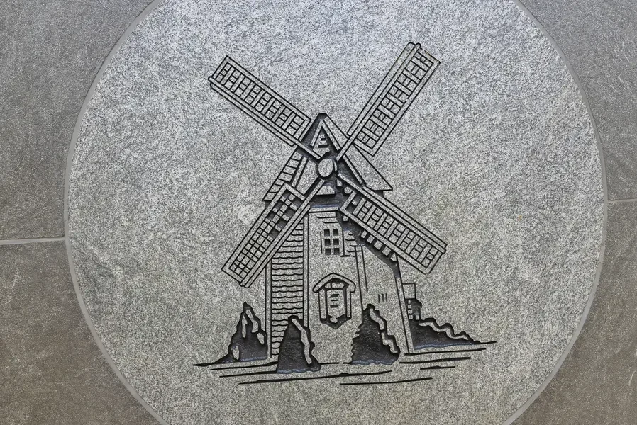 This image shows an intricately carved stone medallion featuring a detailed relief sculpture of a traditional windmill-the logo of Oyster Harbors Golf Club. The circular design is etched into what appears to be light gray granite or similar stone mat