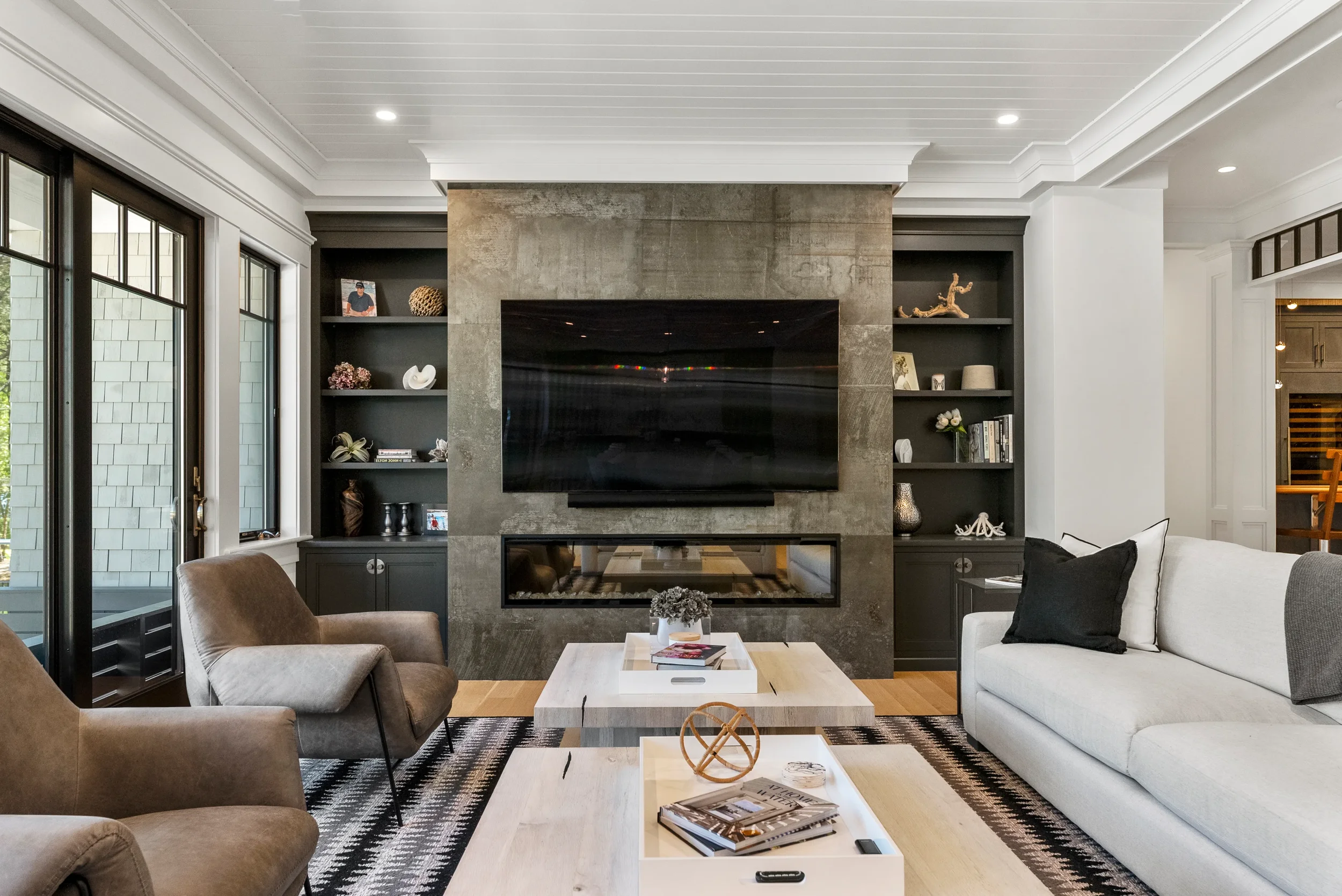 Modern living room with a large flat-screen TV mounted on a gray stone fireplace, surrounded by black shelving units, a white couch with black and gray pillows, two beige armchairs, and a black and white patterned rug.