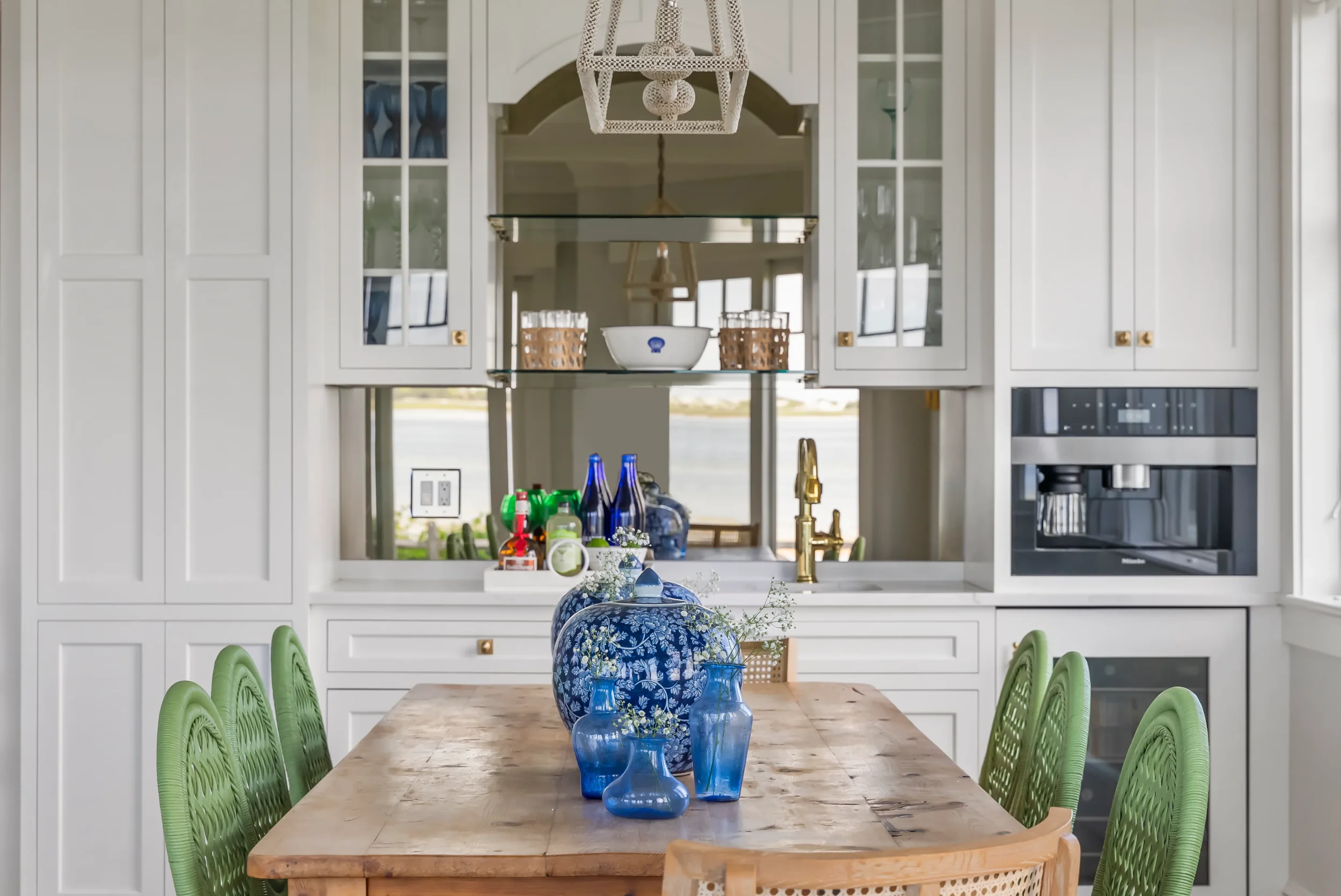 Kitchen with white cabinets, a wooden dining table with green chairs, blue vases, and a window showing a lake outside.