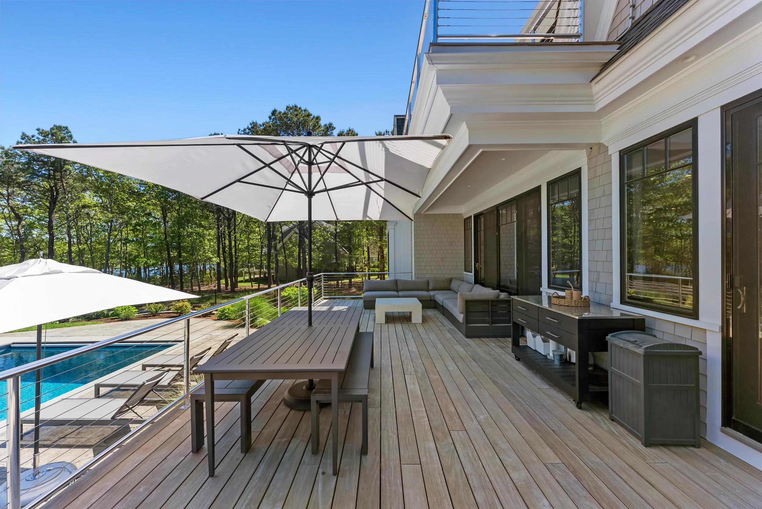 Spacious outdoor patio with wooden decking, large white umbrellas, a dining table with benches, and a seating area with an L-shaped sofa. The patio overlooks a wooded area with trees and a swimming pool.