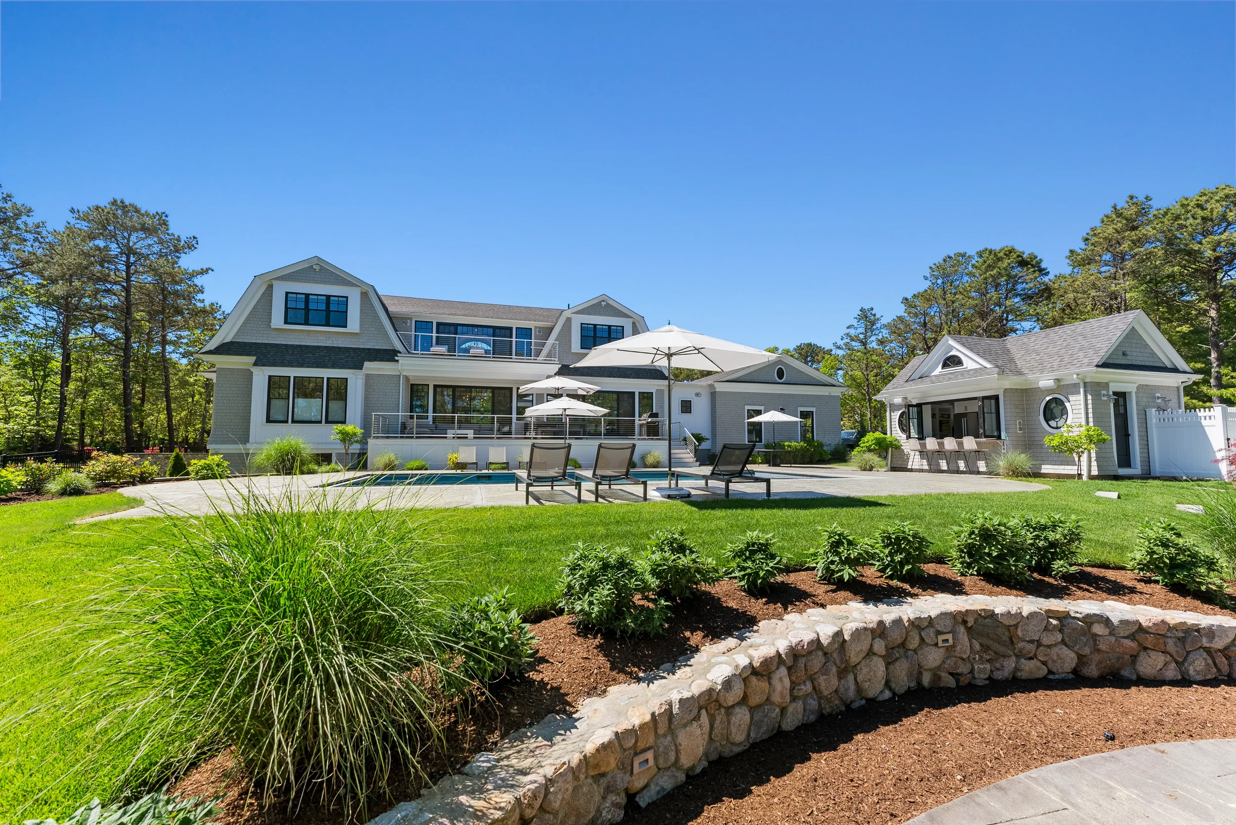 A large backyard with a swimming pool, poolside lounge chairs, umbrellas, and a patio with a bar area, all surrounded by a well-maintained lawn, shrubs, and trees under a clear blue sky.