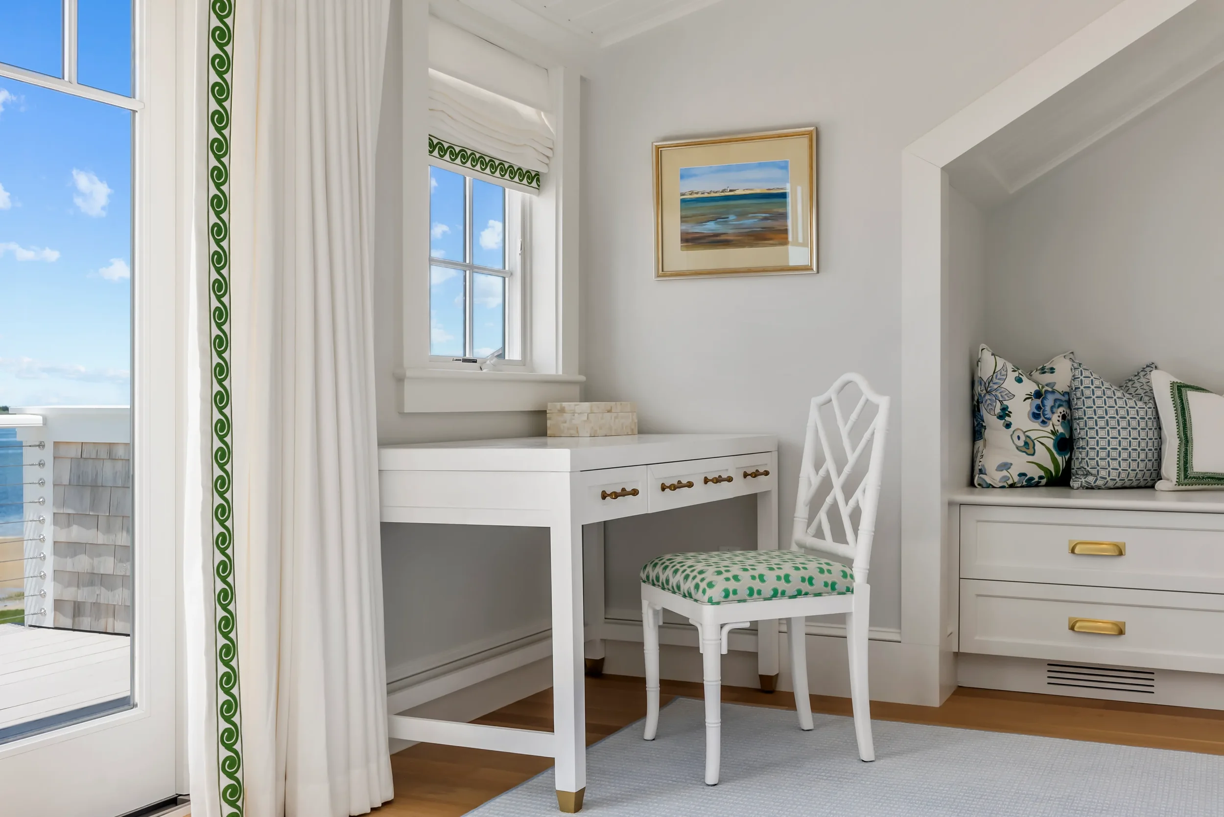 A bright, white corner room with a white desk, a white chair with green polka dot cushion, window with white curtains and green trim, framed landscape painting on the wall, and built-in white bench with decorative pillows.