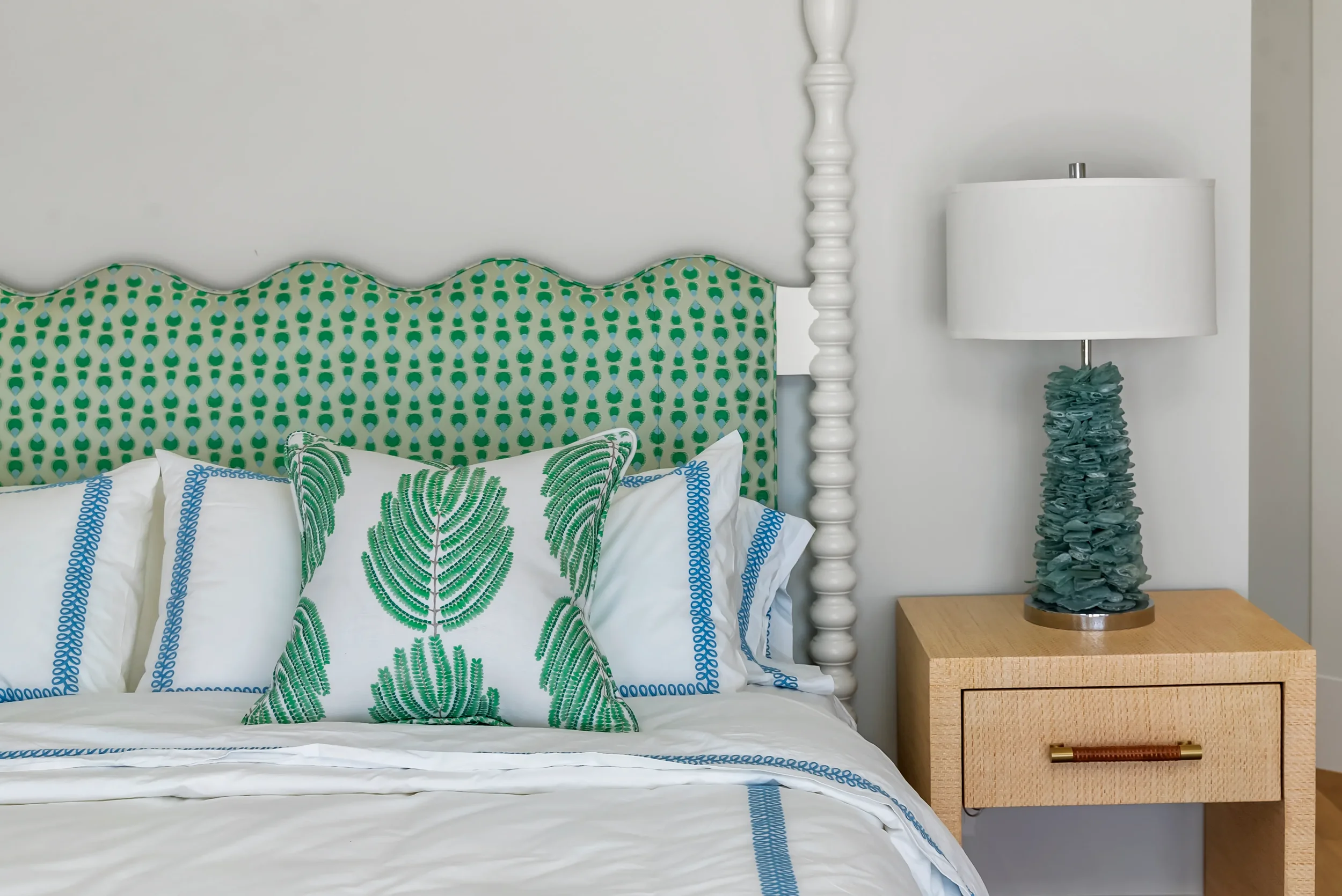 A neatly made bed with white and blue bedding, a green patterned headboard, a decorative pillow with a green fern design, a white pillow with blue accents, a wooden nightstand with a ruffled teal table lamp, in a bright bedroom.