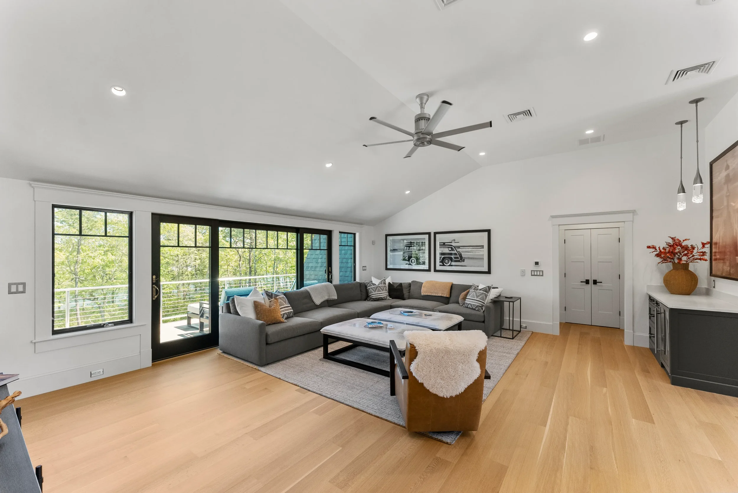 Modern living room with large gray sectional sofa, black and white photographs on the wall, sliding glass door leading to balcony, light wooden flooring, ceiling fan, and decorative elements including pillows, a fur throw, and a vase with red-orange