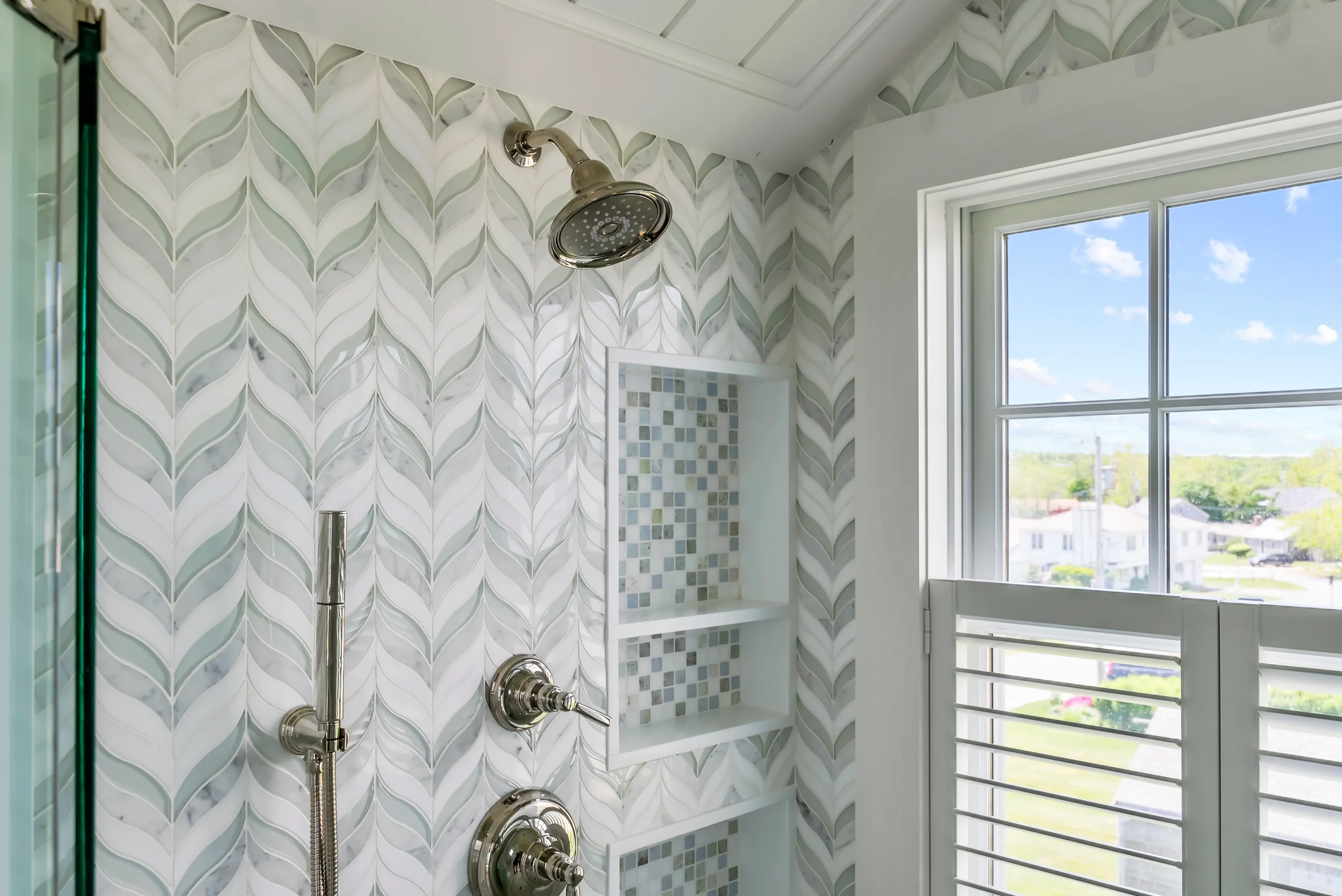 Close-up image of a modern shower with patterned white and grey tile walls, a built-in tiled niche, a chrome showerhead, and a window showing a cityscape and blue sky.
