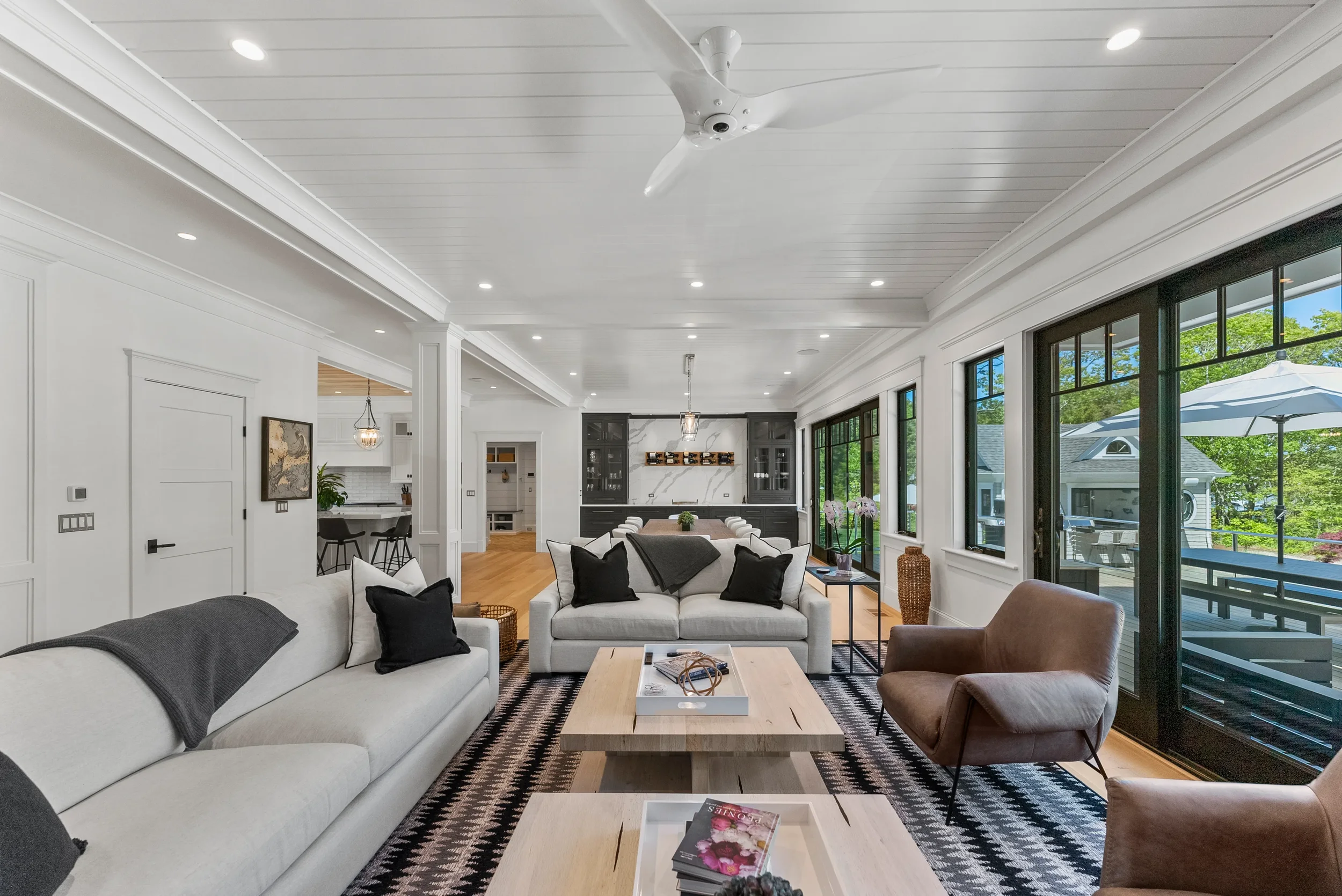 Bright living room with white walls, large windows, and modern furniture, including a white sofa with black and white pillows, a brown armchair, wooden coffee tables, and a black and white patterned rug, overlooking a patio with outdoor furniture and