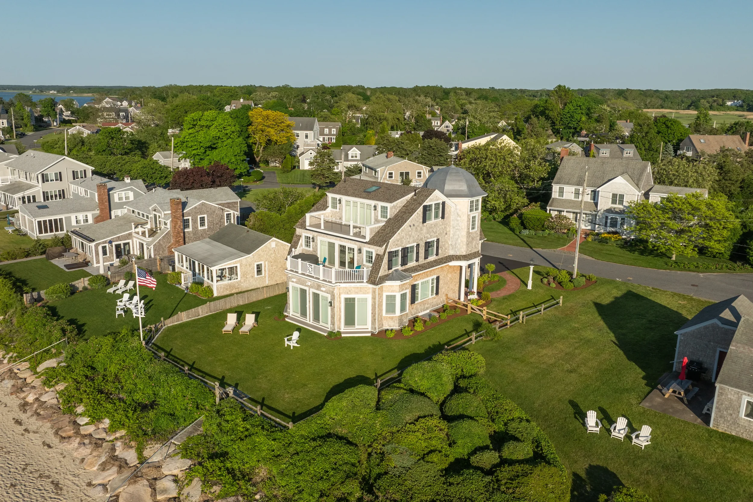 Aerial view of a coastal neighborhood with large houses, green lawns, trees, and a rocky shoreline along the water.