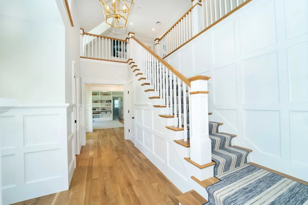 Bright foyer with wooden stairs, white panel walls, and hardwood flooring, leading to a room with built-in shelves.