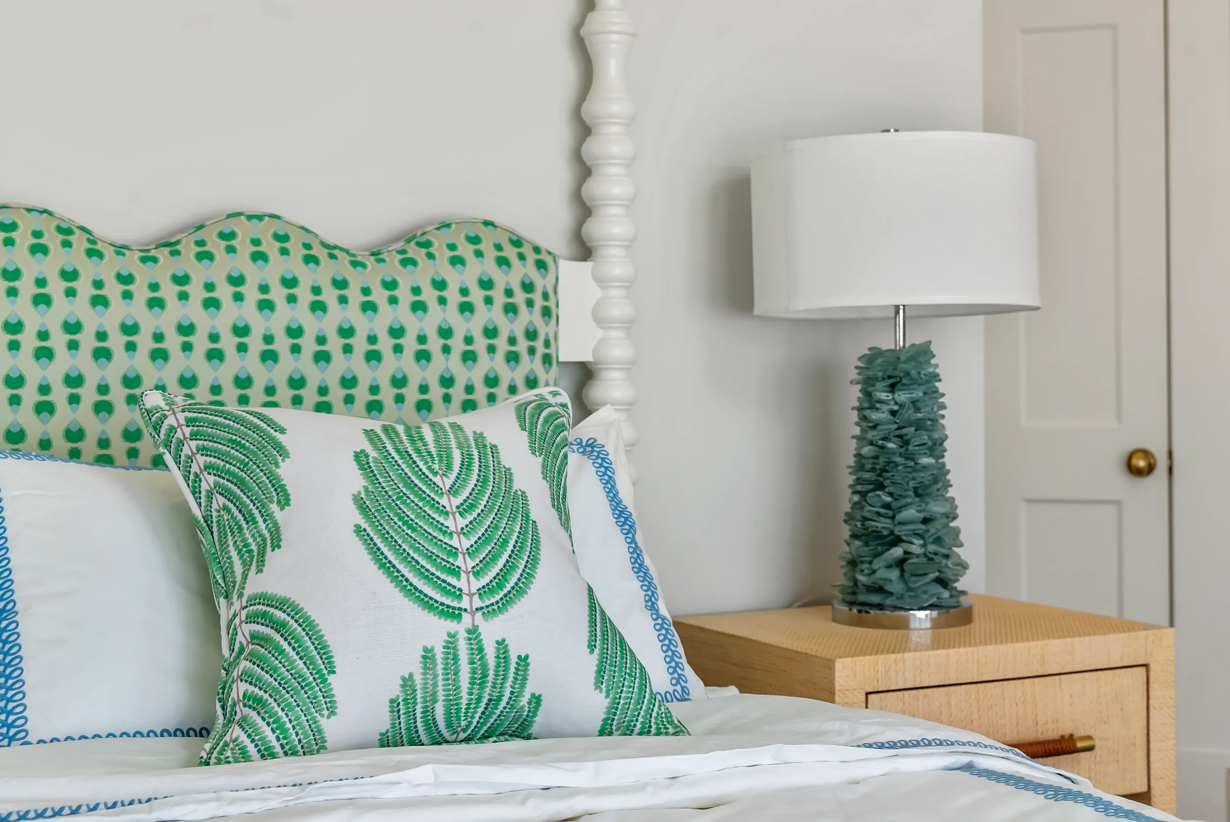 A bed with white bedding, green and white pillows with leaf patterns, a wooden nightstand, a ceramic lamp with a white shade, and a door in a white wall.