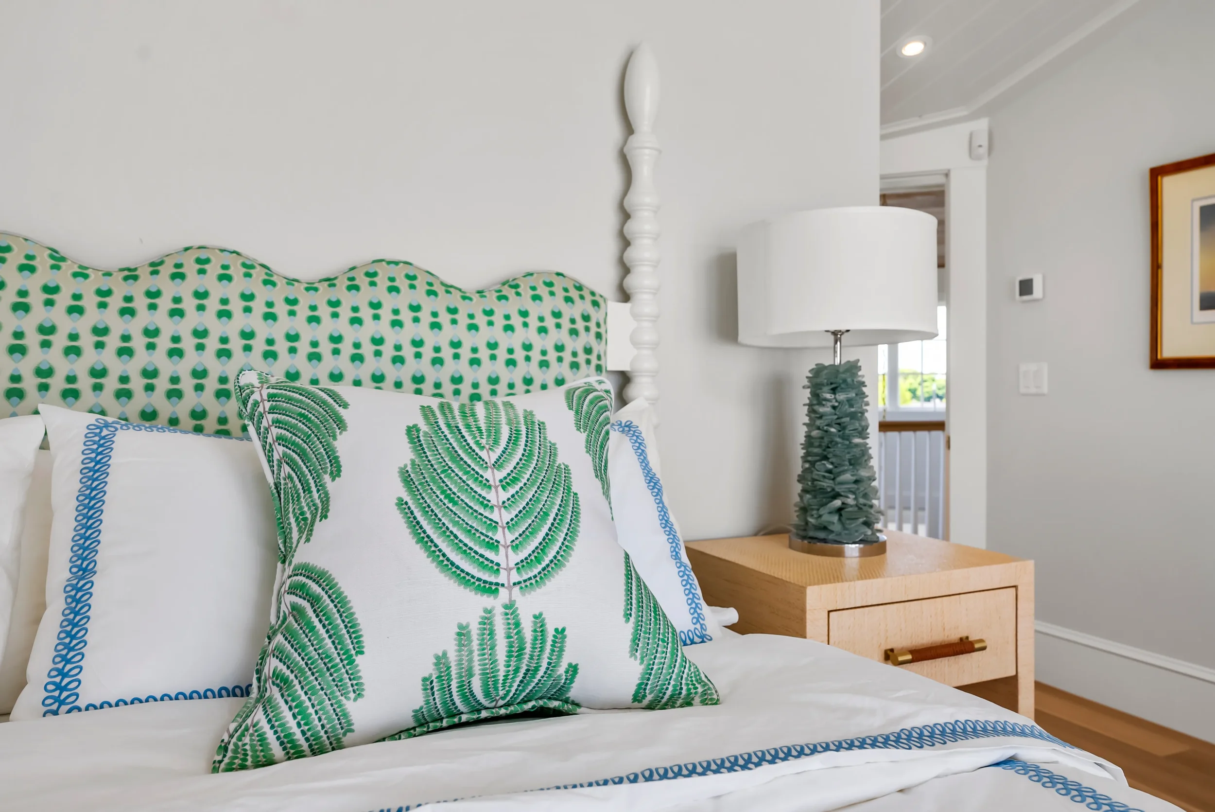 A bedroom interior with a white bed, decorative pillows with green fern and blue leaf patterns, a green and white patterned headboard, and a wooden nightstand with a modern lamp that has a textured base and white shade.