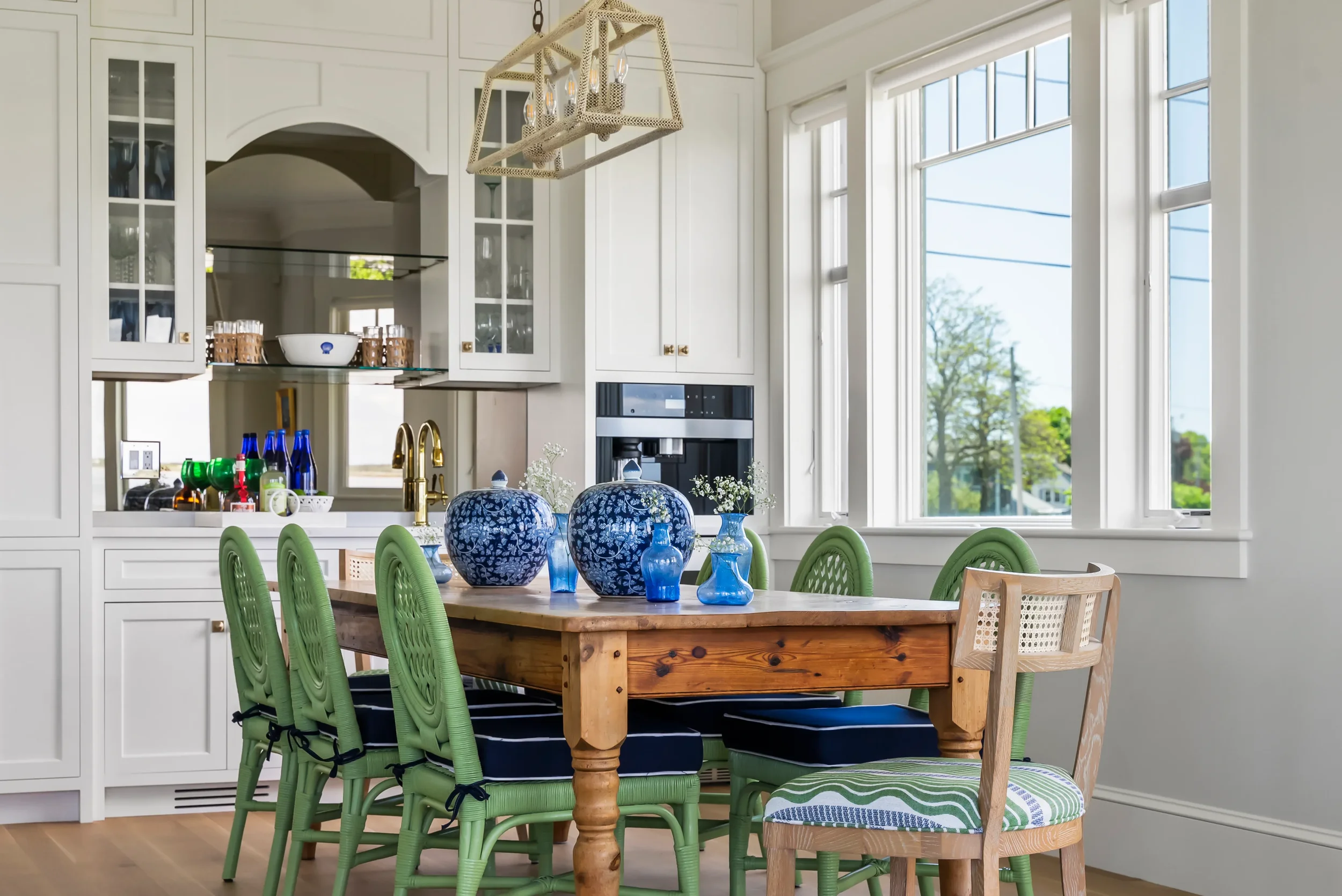 A dining area with a wooden table topped with blue and white vases, surrounded by six green chairs with black cushions and one wooden chair with a patterned fabric. The room has large windows, white cabinets, and a light-colored floor.